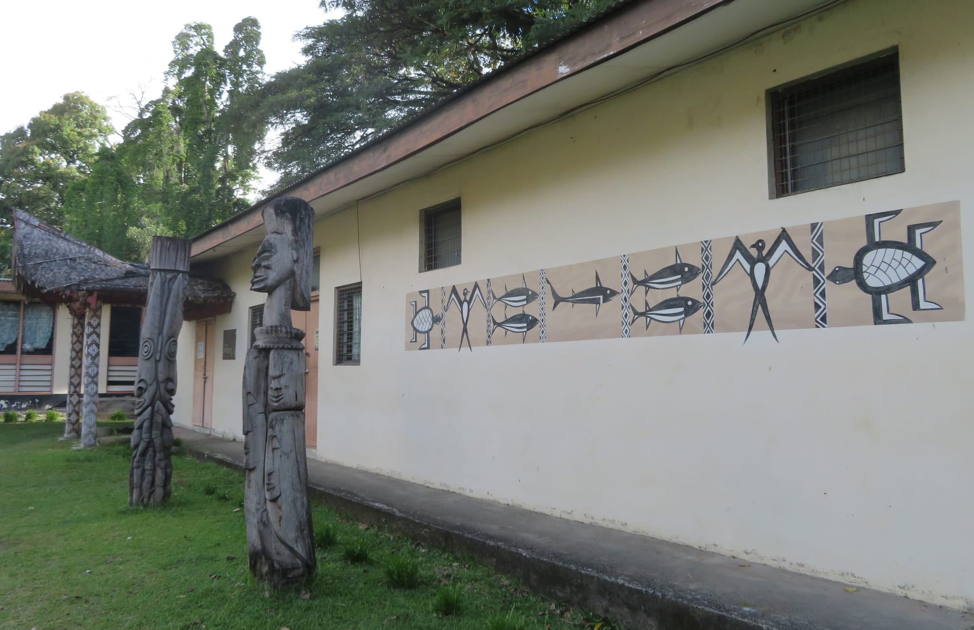 Two tall carved wooden posts with stylised faces stand on a lawn beside a low cream building whose wall is painted with a black and white frieze of fish, birds and a turtle, with a thatched shelter and tall trees behind