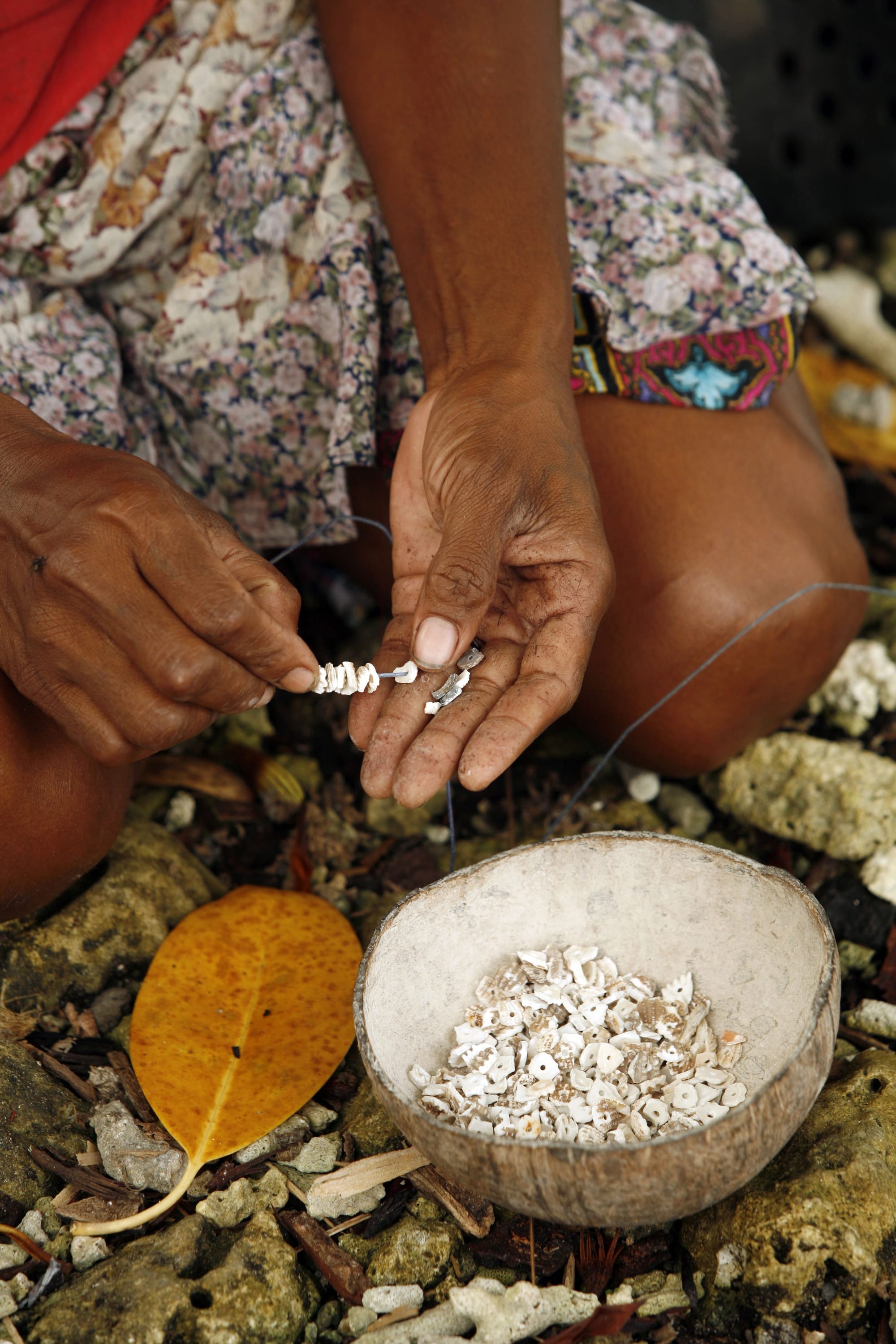 A pair of hands threading small white shell discs onto a thin line, above a coconut-shell bowl filled with more drilled discs on a coral-strewn ground