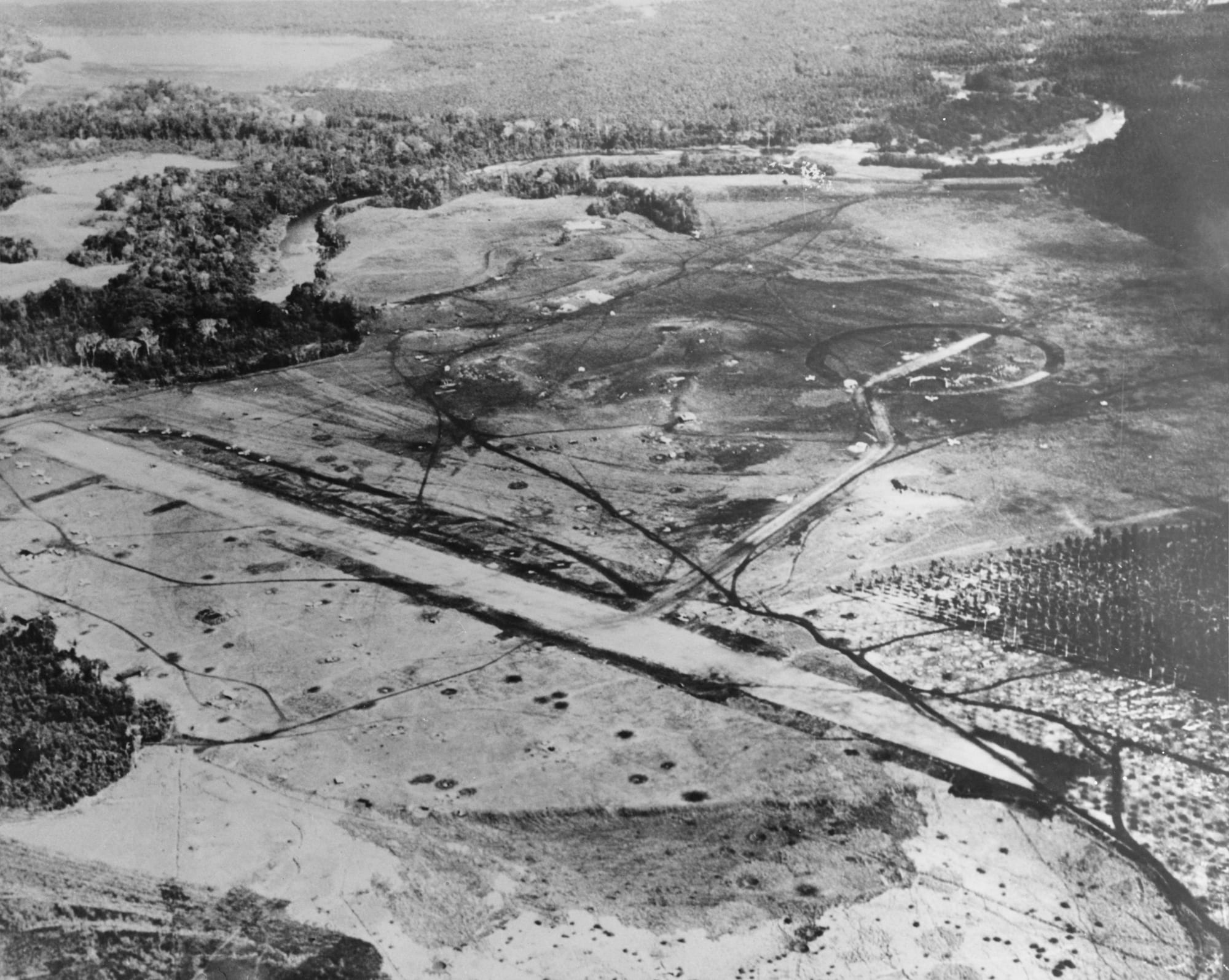 An aerial photograph of a cleared airstrip with dispersal tracks and bomb craters, ringed by jungle and a river