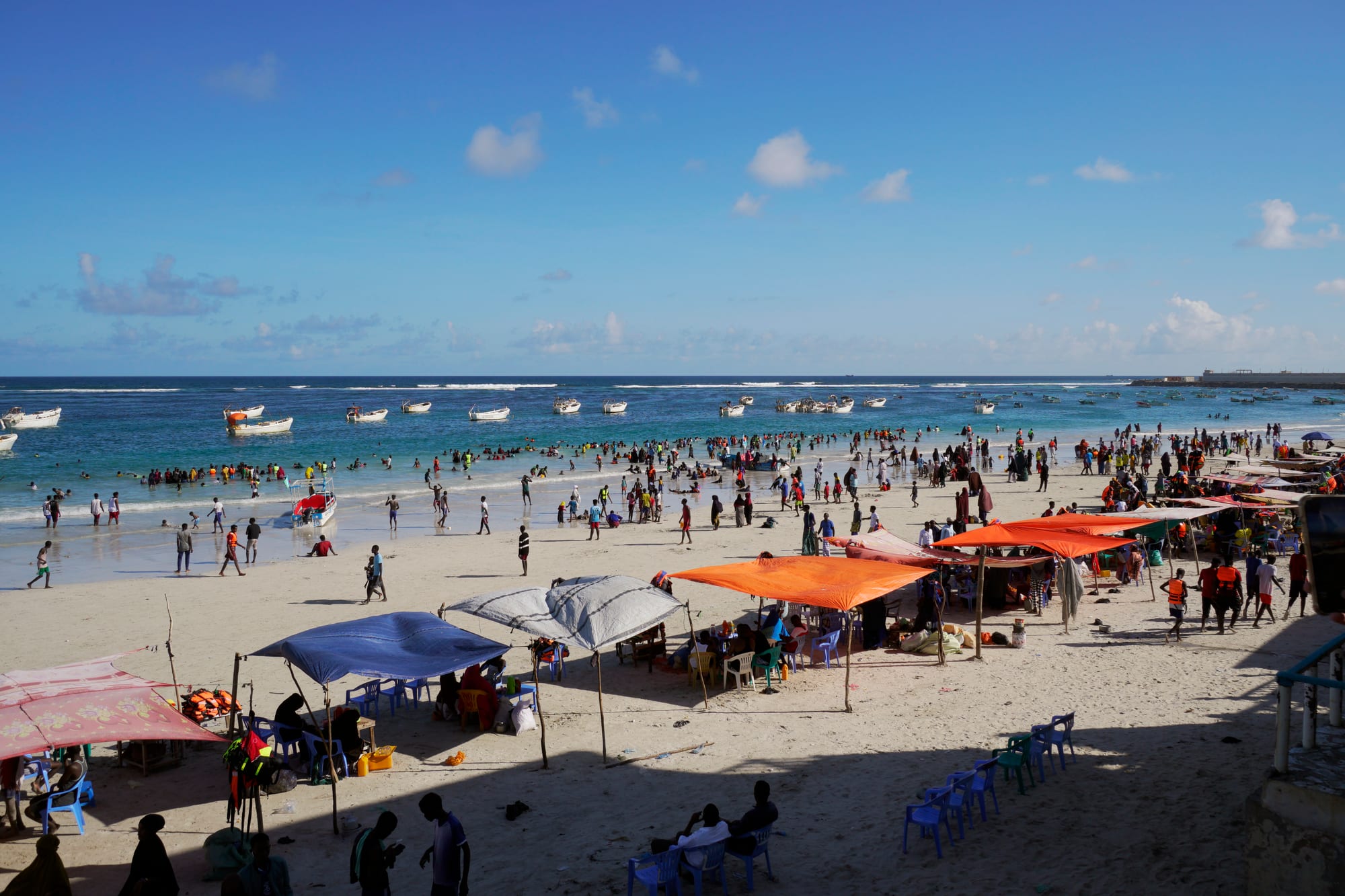A crowded white-sand beach with fishing boats offshore and sunshades on the sand
