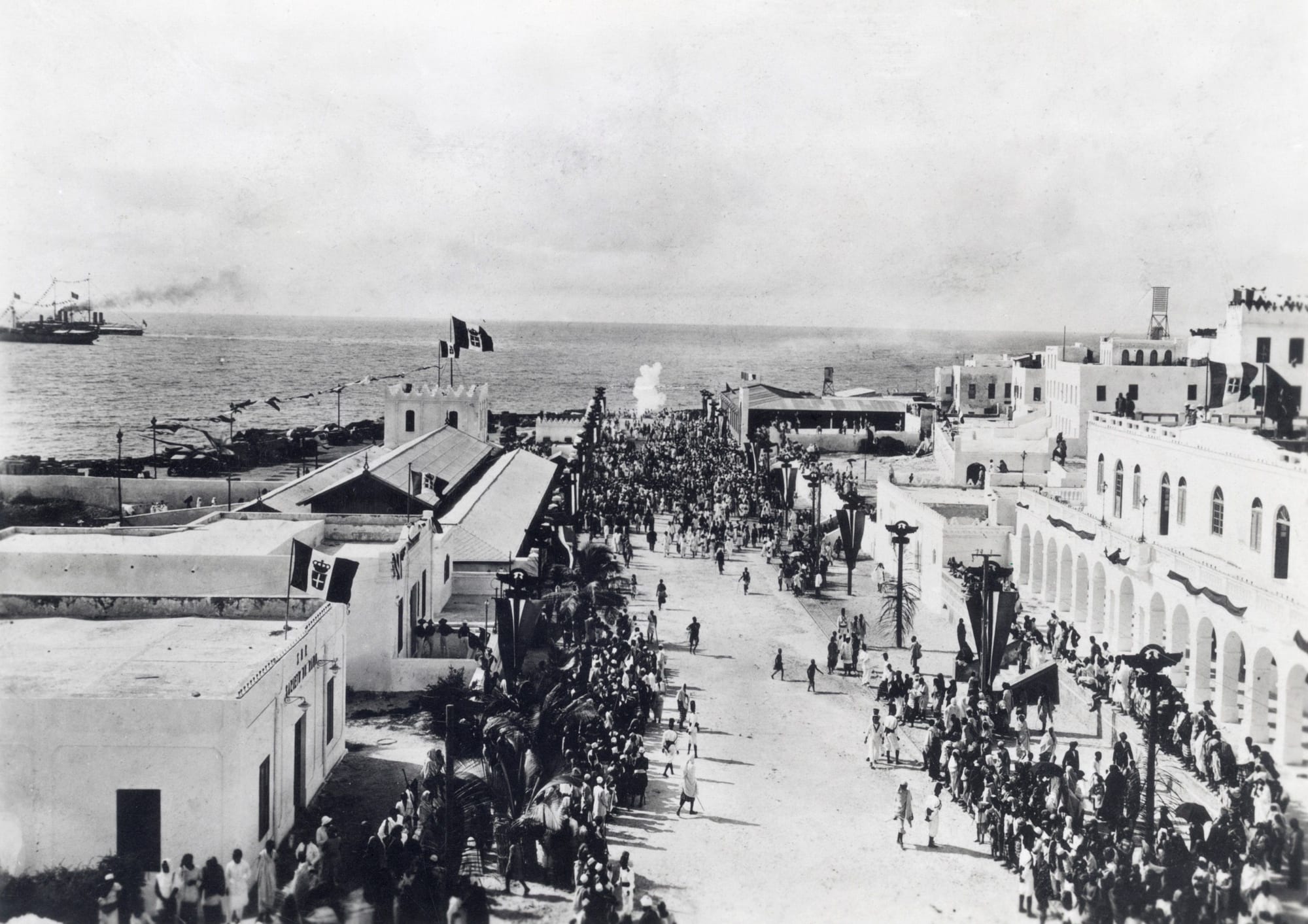 A black-and-white 1925 photograph looking down a wide street in Mogadishu lined with white arcaded buildings, flags and a large crowd, leading to the sea where a steamship lies offshore
