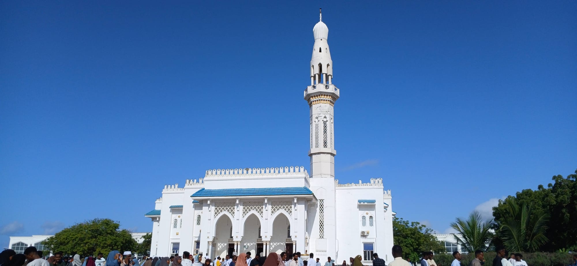 A large white mosque with a tall slender minaret, arched entrances and blue-trimmed roofs under a clear blue sky, with a crowd of people gathered in front