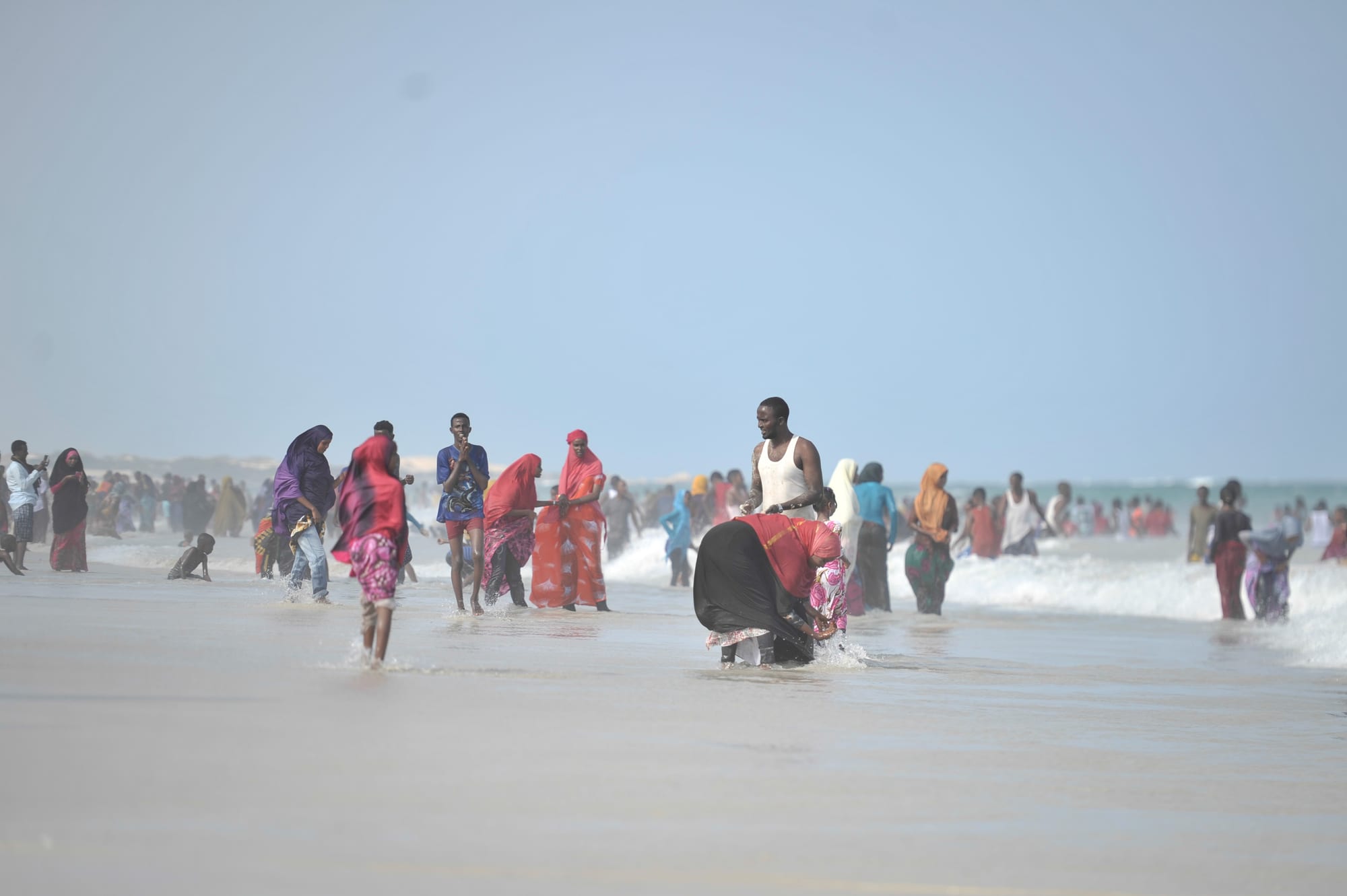 Crowds of people, many women in brightly coloured headscarves and wraps, wade and walk in shallow surf along a wide beach under a hazy sky