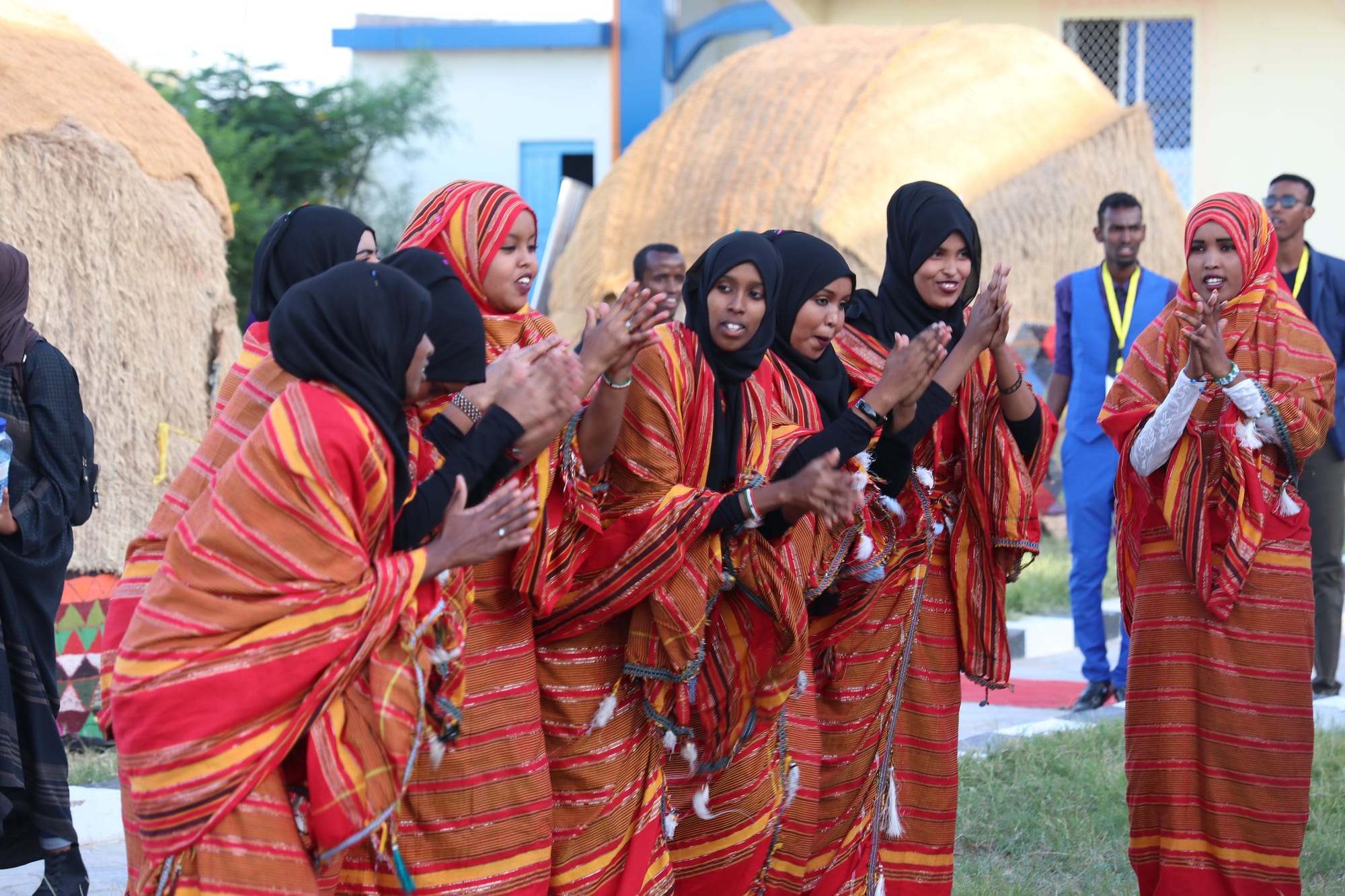 A group of young women in red and orange striped wraps and black headscarves clap and sing in a line, in front of two large domed huts covered with woven grass