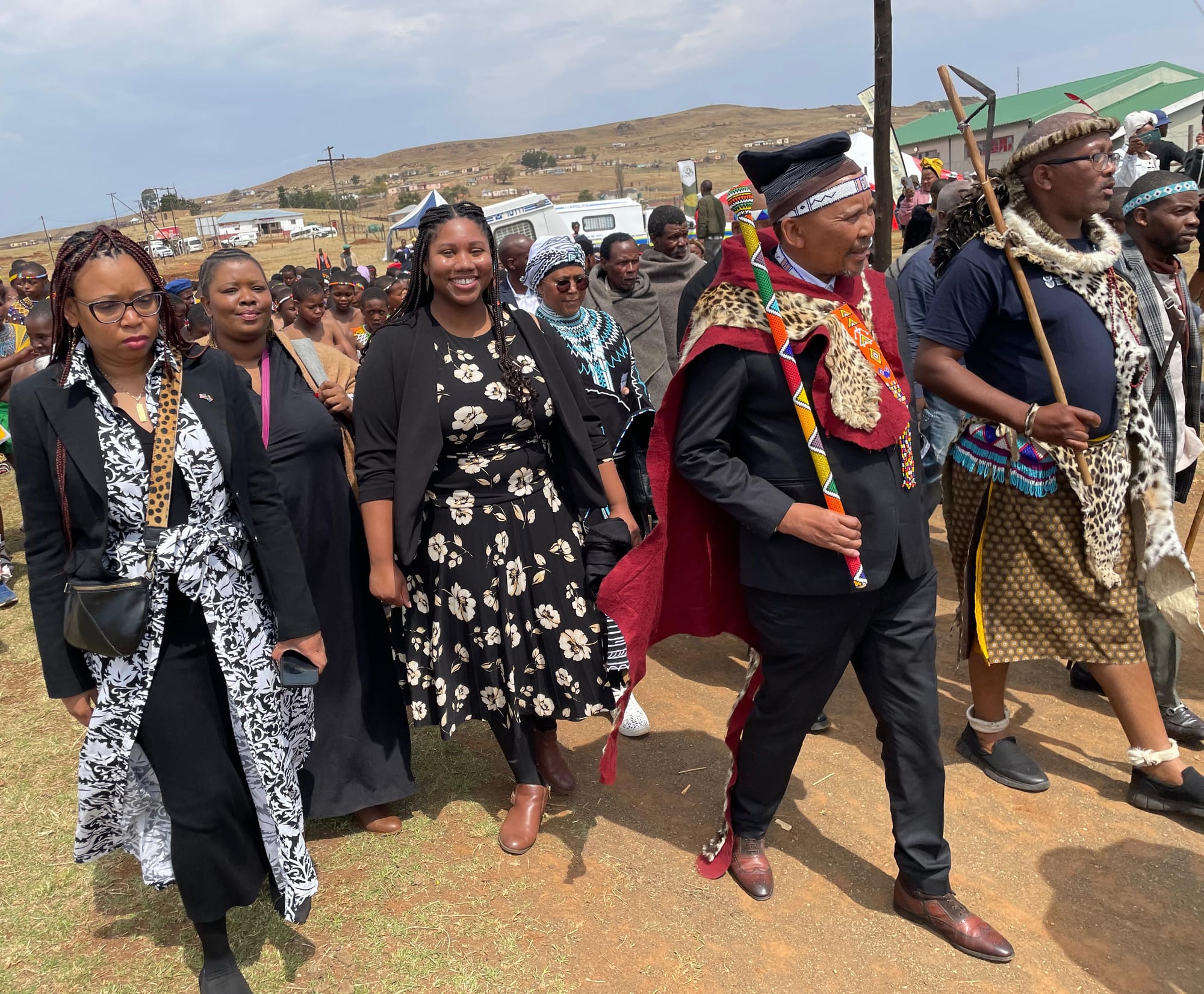 A group of people walking together across open ground, some in everyday clothes and some in traditional dress including leopard-print capes, beaded sashes and headbands, with one man carrying a long staff