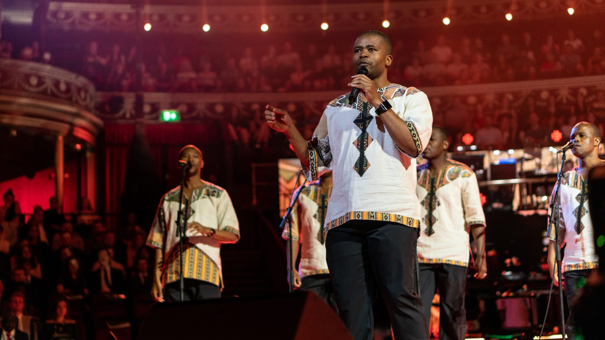 A male singer in a white shirt with a patterned border sings into a handheld microphone on a stage, with other singers in matching shirts standing at microphones behind him and an audience in tiered seating beyond