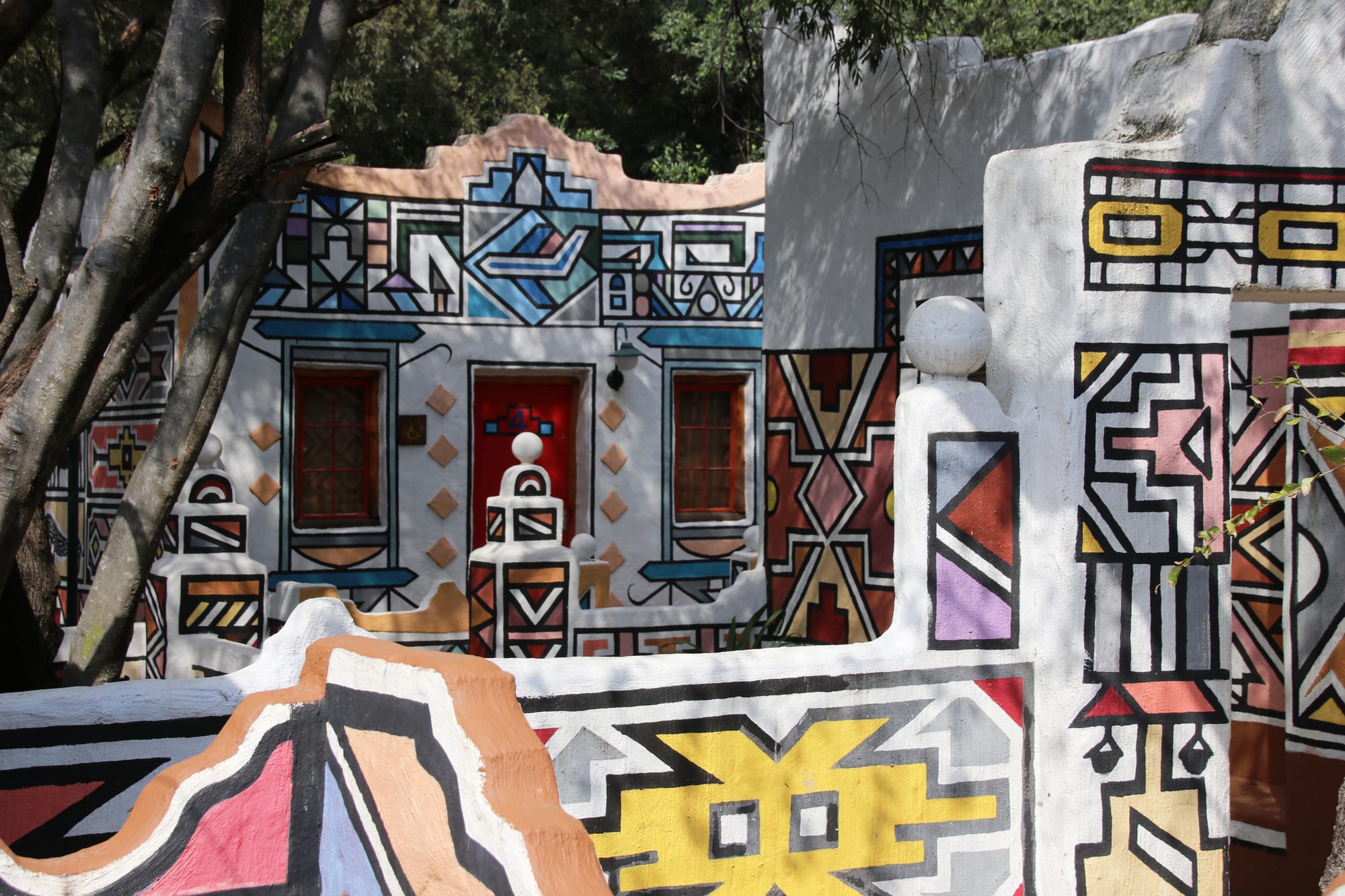 Whitewashed walls and a low building painted with bold geometric patterns in black outline, filled with blue, yellow, pink and red, around a red door and two windows