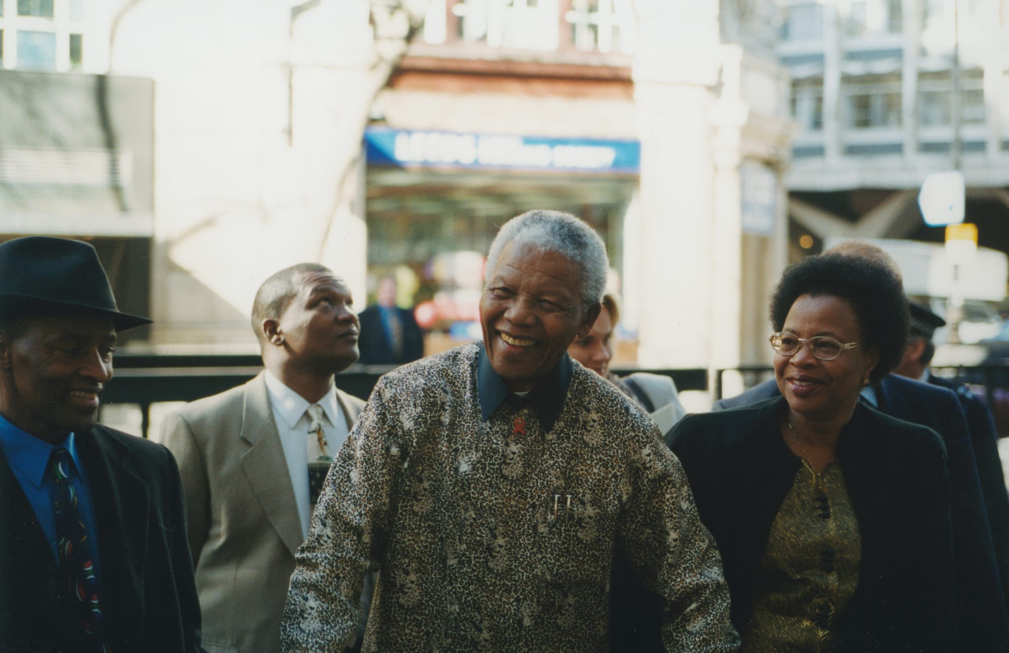 A smiling Nelson Mandela in a patterned shirt walking among a small group of people on a city street