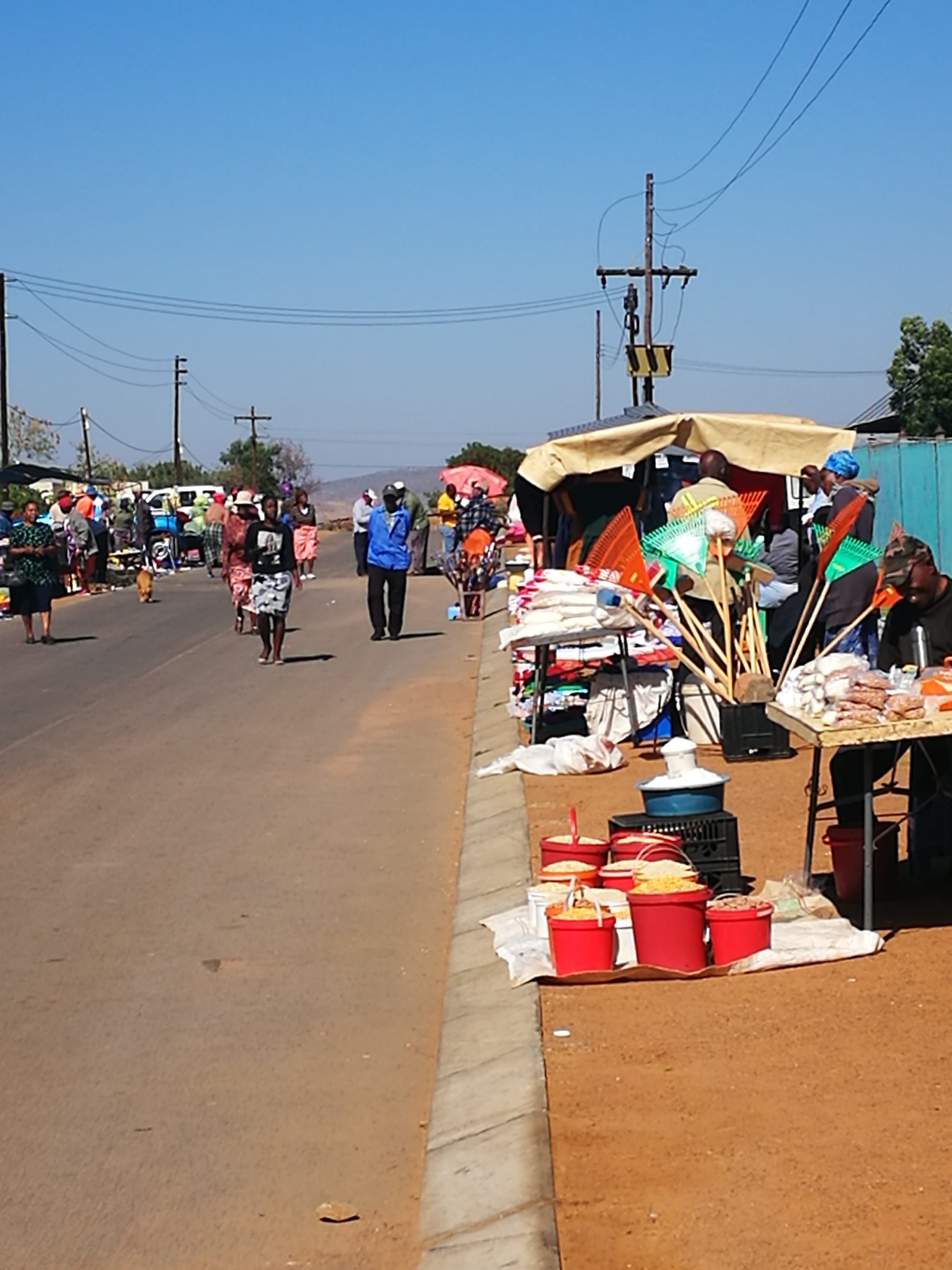Vendors and customers at an open-air street market selling clothes, buckets of grain and household goods