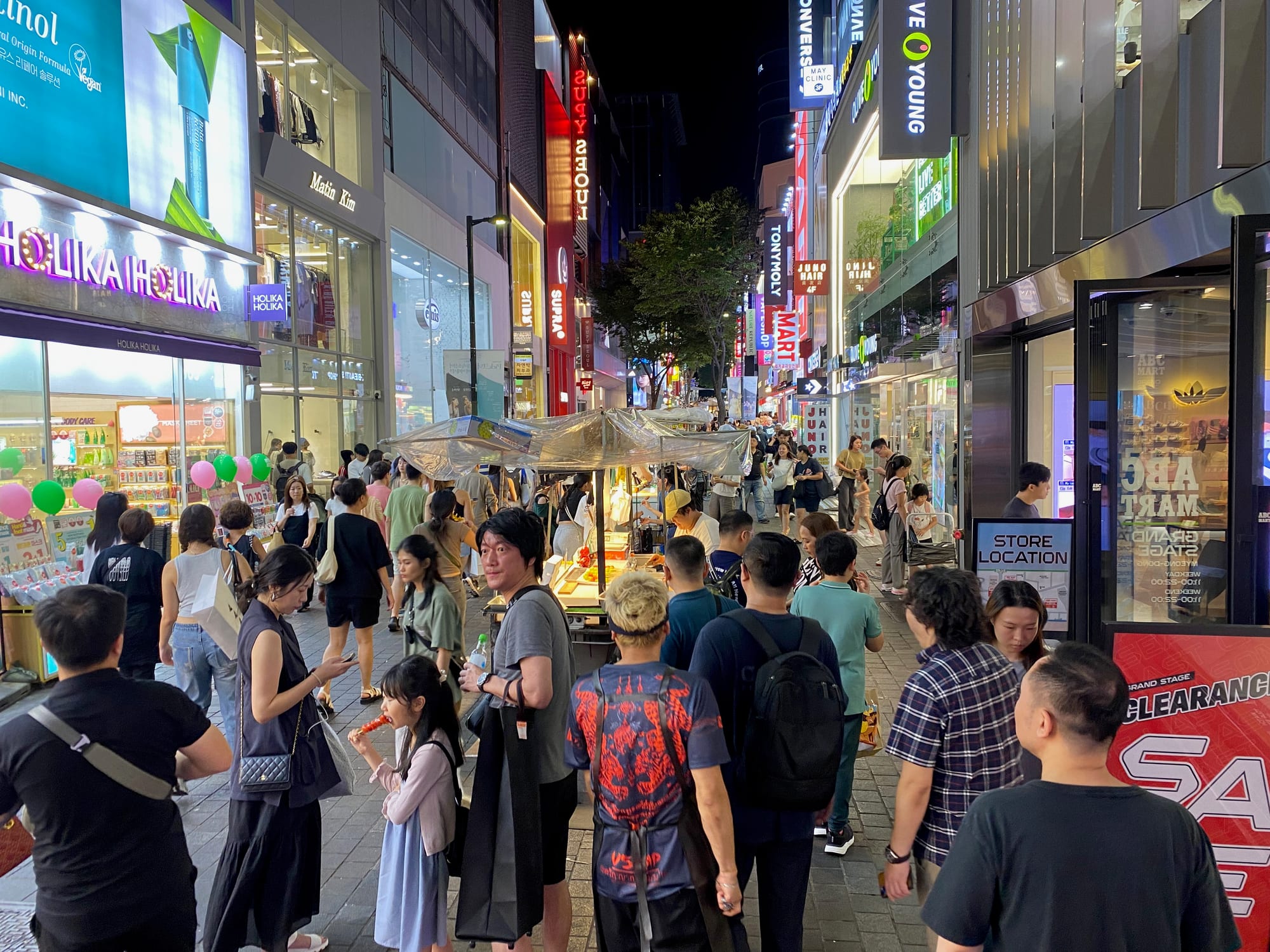 A crowded Seoul shopping street lit by neon storefront signs after dark, with vendors and pedestrians filling the road