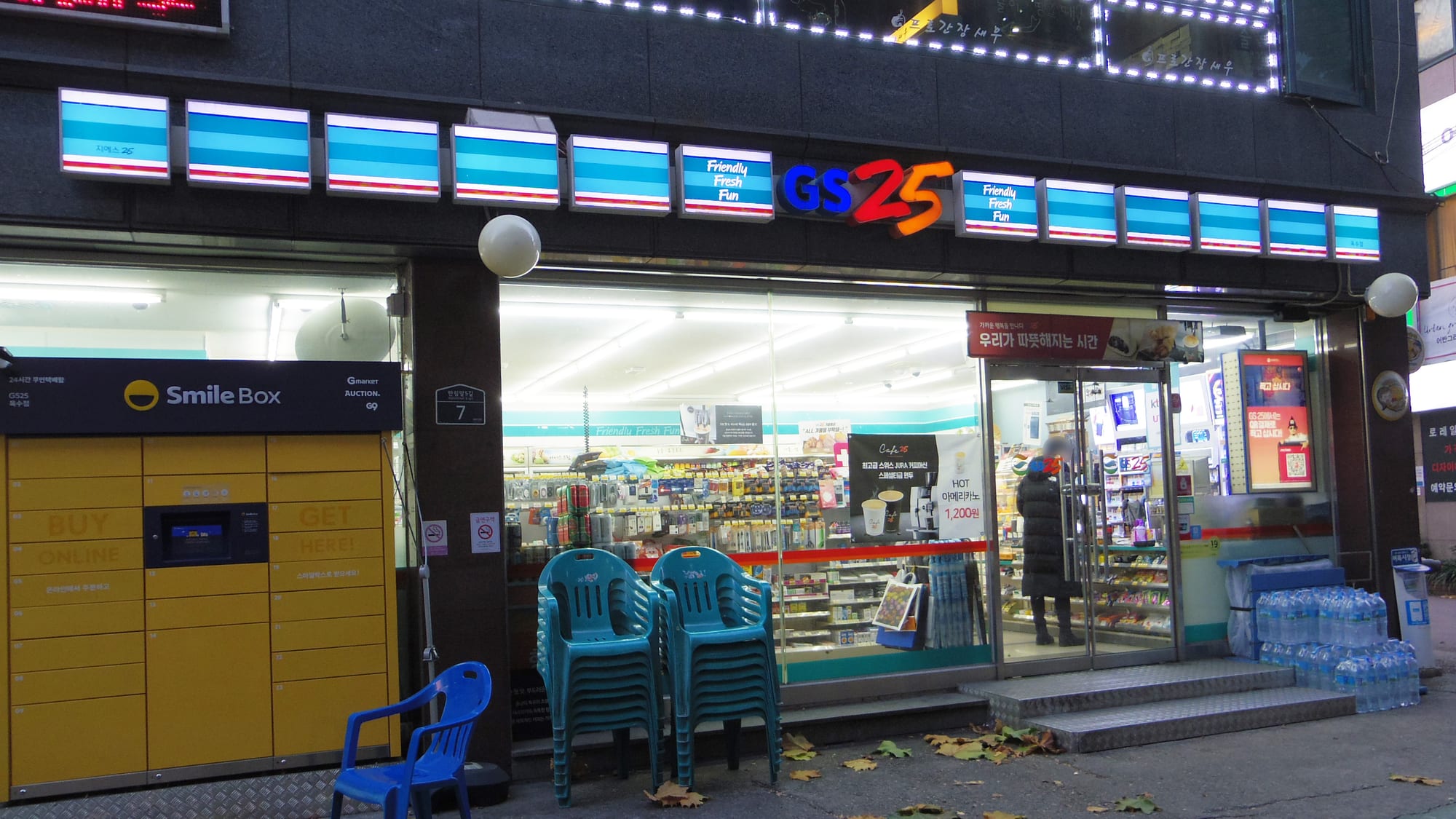 The brightly lit storefront of a GS25 convenience store at dusk, with plastic chairs and a parcel locker outside and a customer visible inside