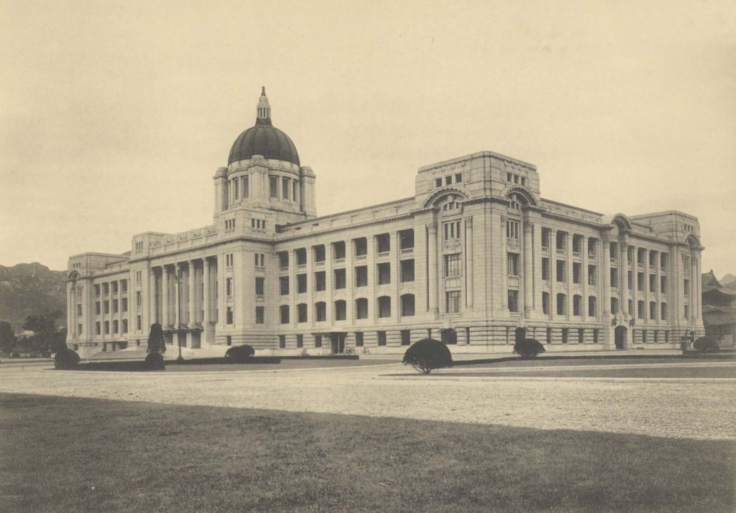 A sepia photograph of a long stone government building with a central dome, seen across an open forecourt
