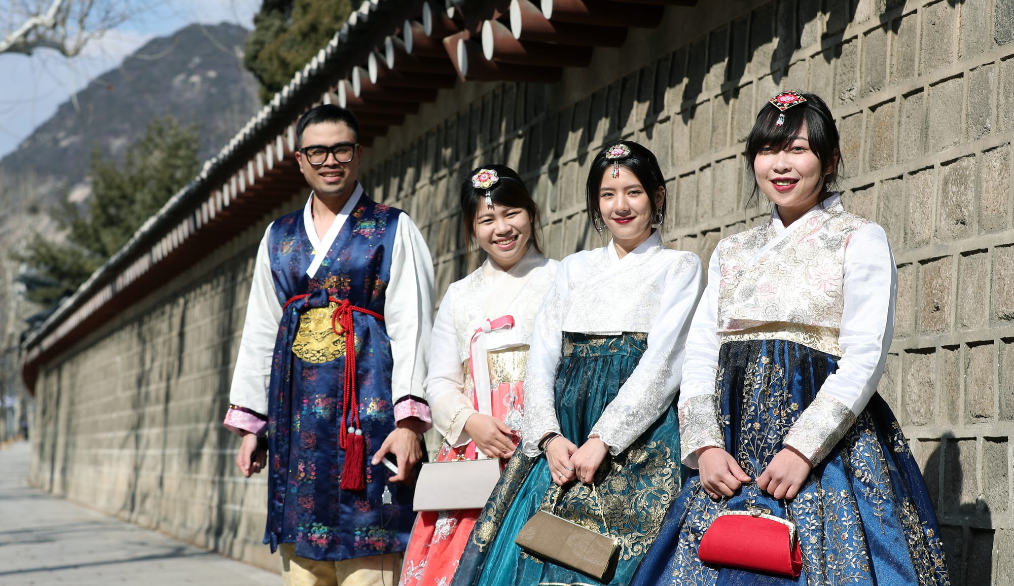 A man and three women in hanbok stand smiling beside a long stone wall topped with a tiled roof, the women in embroidered jackets and full skirts of pink, teal and deep blue