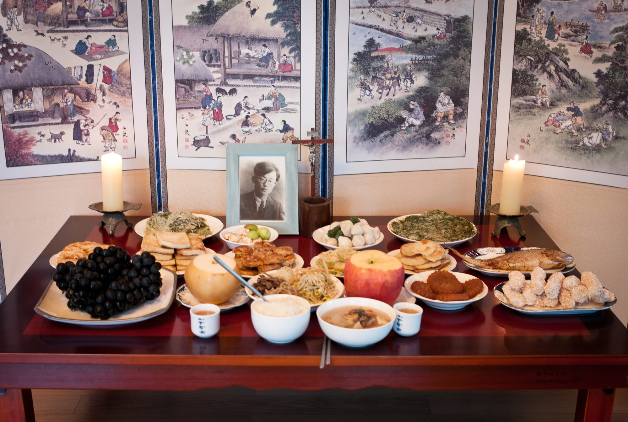 A dark wooden table laid with plates of fruit, rice cakes, fried fish, pancakes and bowls of soup and rice, with small cups, two lit candles and a framed black and white portrait, set against painted folding screens
