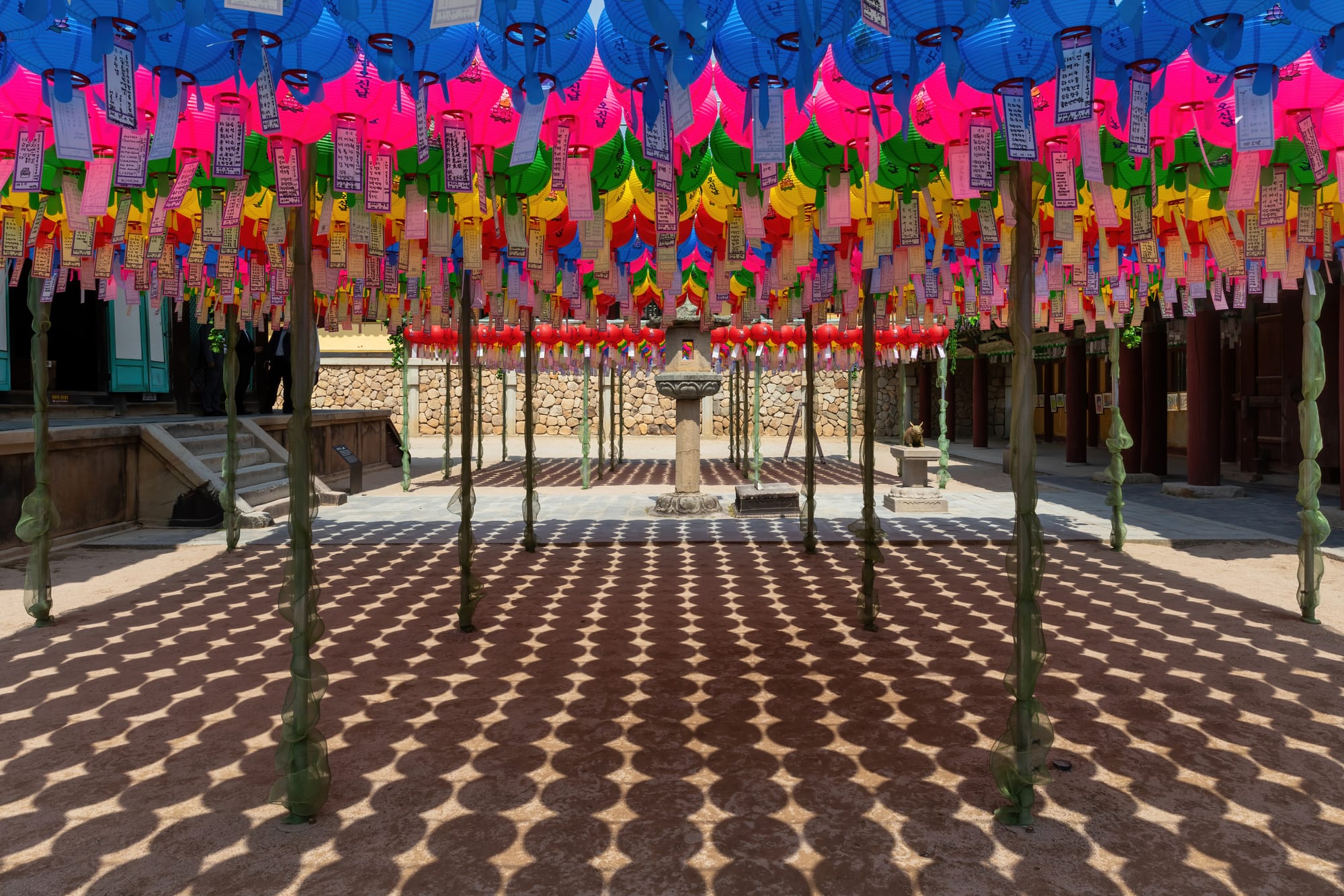 Rows of pink, blue, green and yellow paper lanterns with hanging paper tags fill the air above a temple courtyard, casting patterned shadows on the paving below