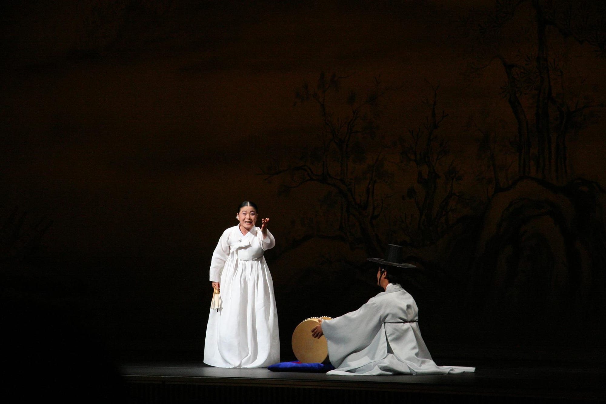 A woman in a white hanbok stands on a dark stage holding a folded fan and gesturing as she sings, while a man in a white robe and a black brimmed hat kneels beside a barrel drum