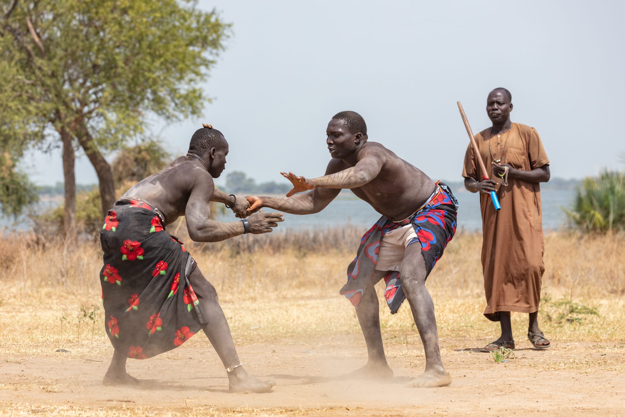 Two bare-chested wrestlers in patterned wraps crouch and reach towards each other on dusty ground, while a man in a long brown robe holding a stick watches beside them, with a river behind