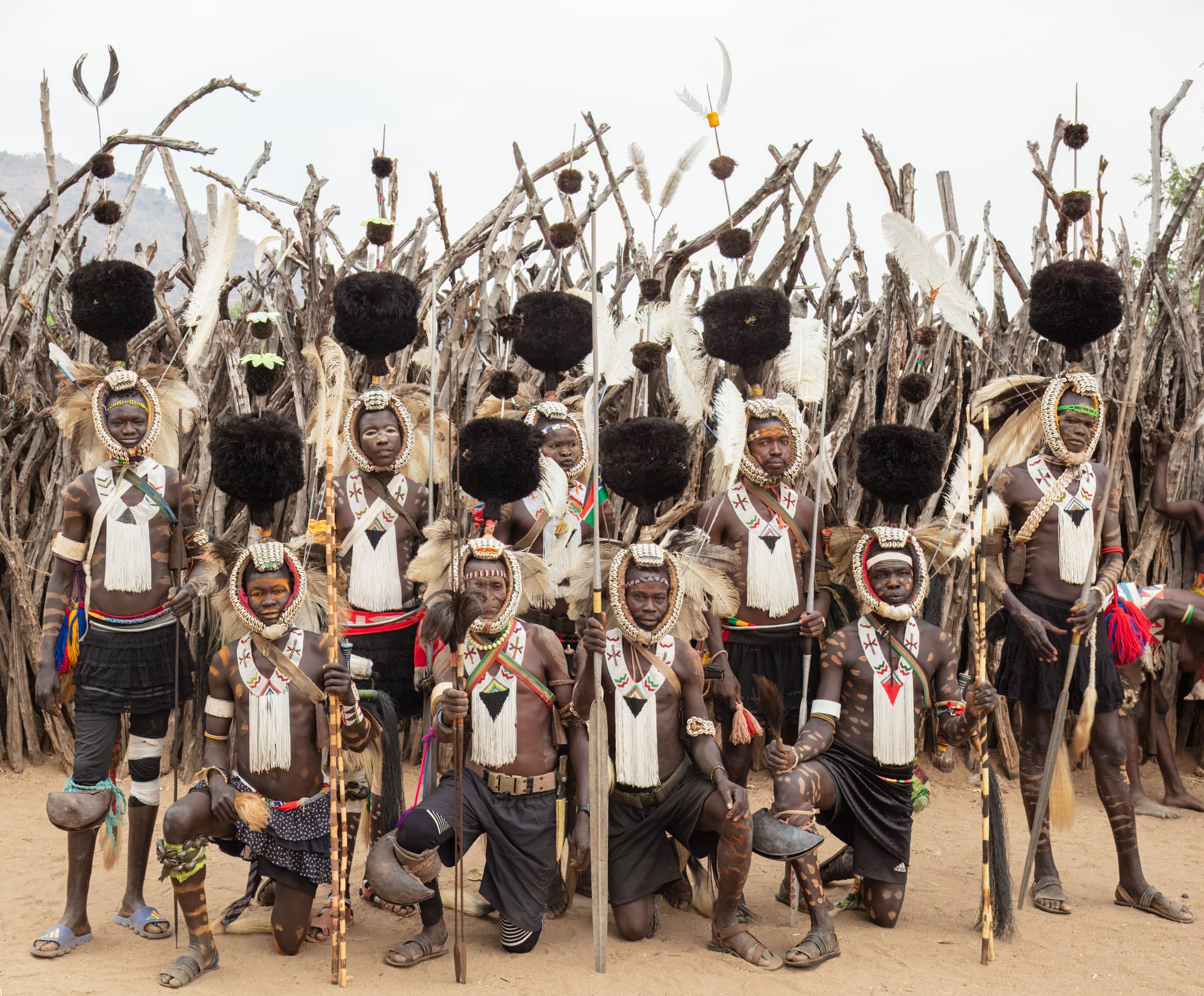 A group of men, some standing and some kneeling, wear tall black plumed headdresses, cowrie-shell headbands and white beaded chest ornaments, with painted markings on their bodies, in front of a fence of wooden branches