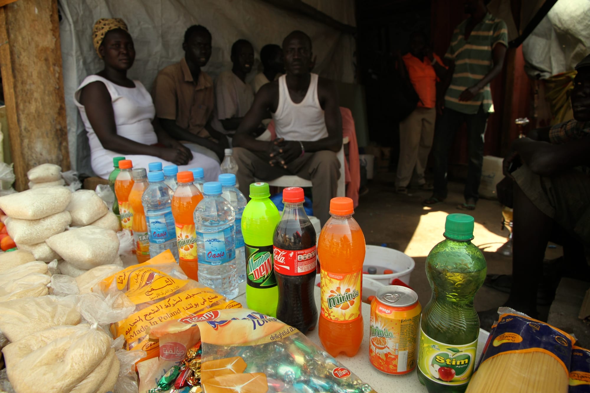 A small market stall selling bottled drinks, rice and other goods, with several people sitting nearby
