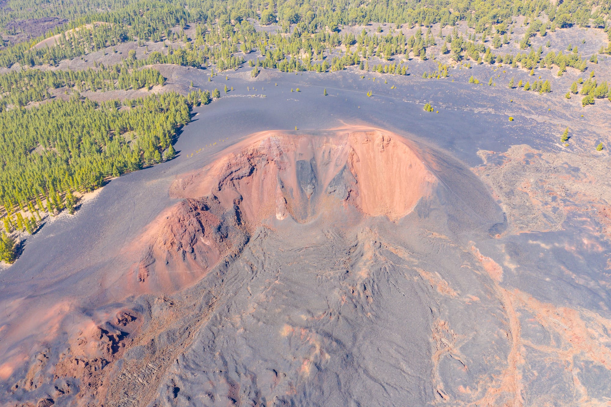 An aerial view of a reddish volcanic crater surrounded by pine forest