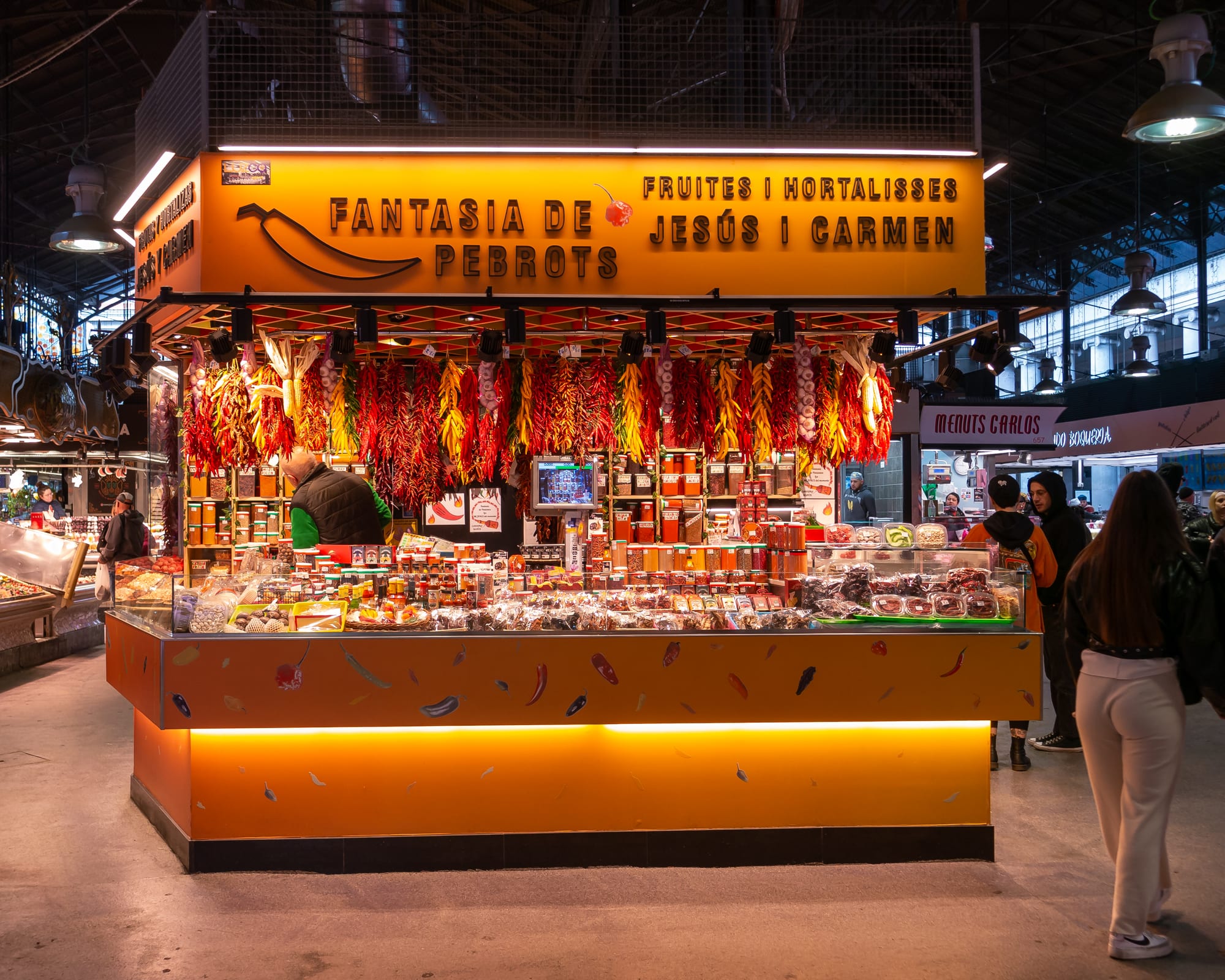 A busy covered-market food stall hung with strings of dried peppers, with shoppers browsing produce and spices in front