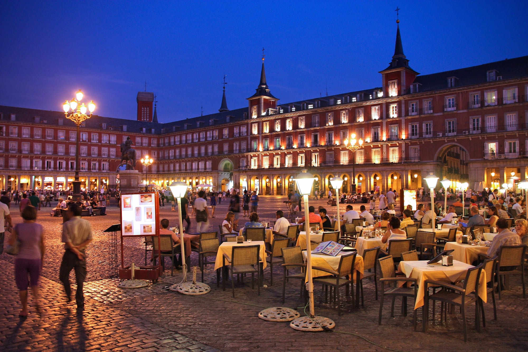 Rows of outdoor café tables filled with diners in Madrid's Plaza Mayor at dusk, with lamplight and a historic red facade behind