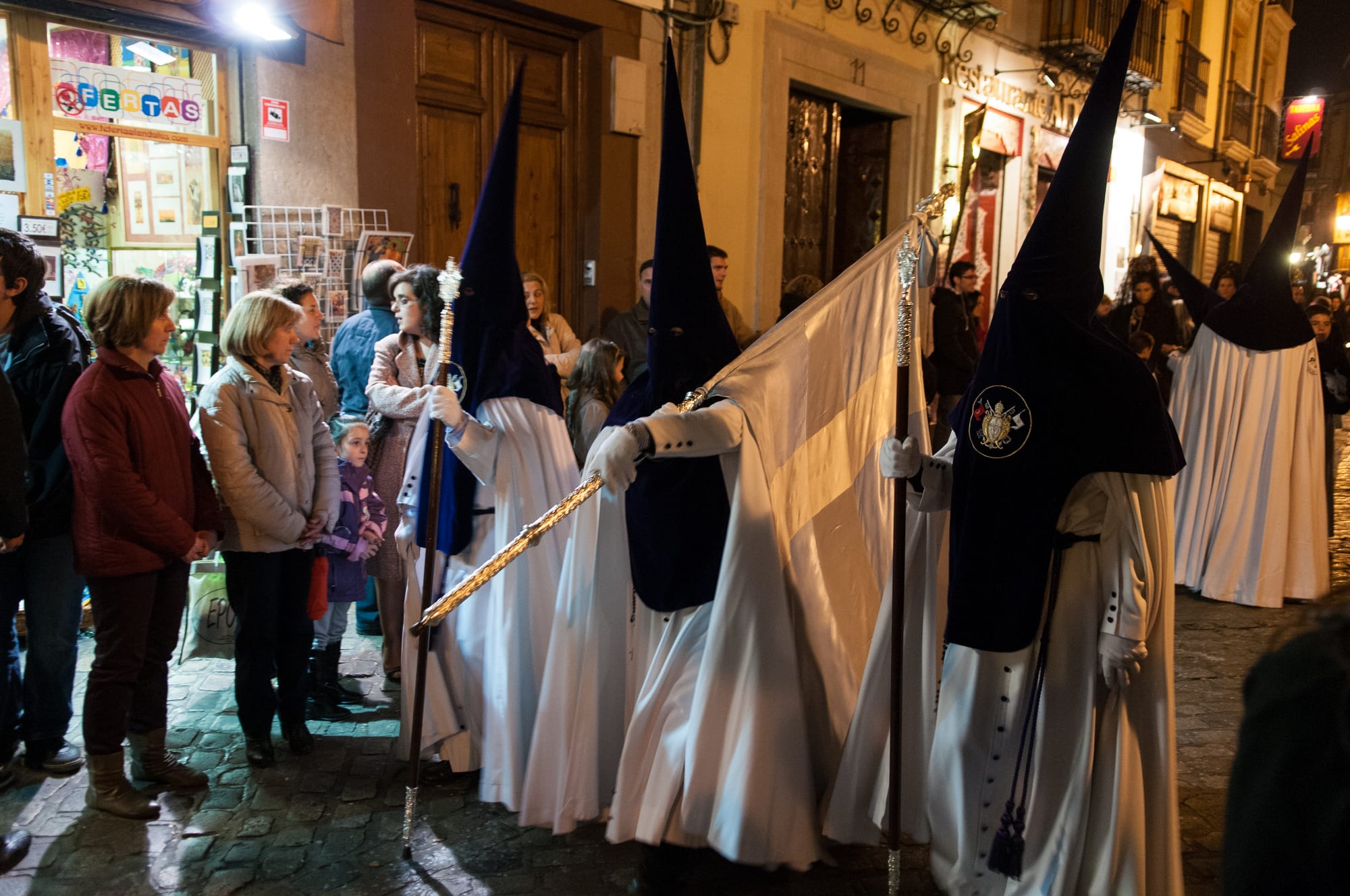 Penitents in long white robes and tall pointed dark blue hoods carry a white banner and silver staffs along a narrow street at night, watched by people standing in shop doorways