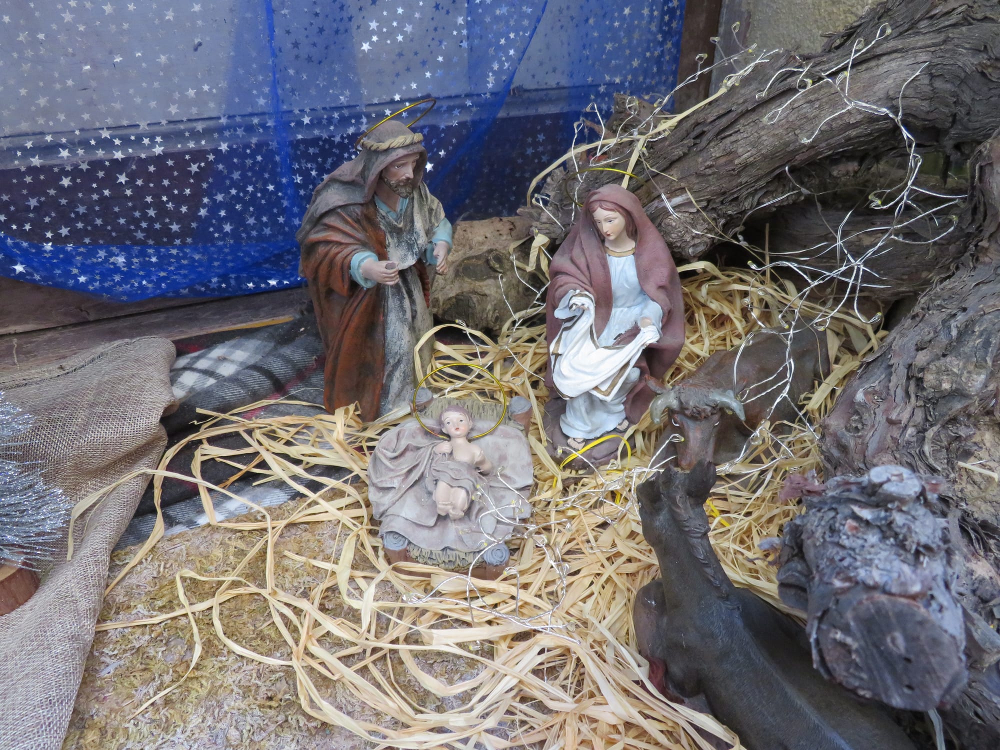Painted figures of Mary and Joseph kneel beside the infant Jesus lying on straw, with animal figures nearby, set among pieces of old wood beneath a starry blue cloth