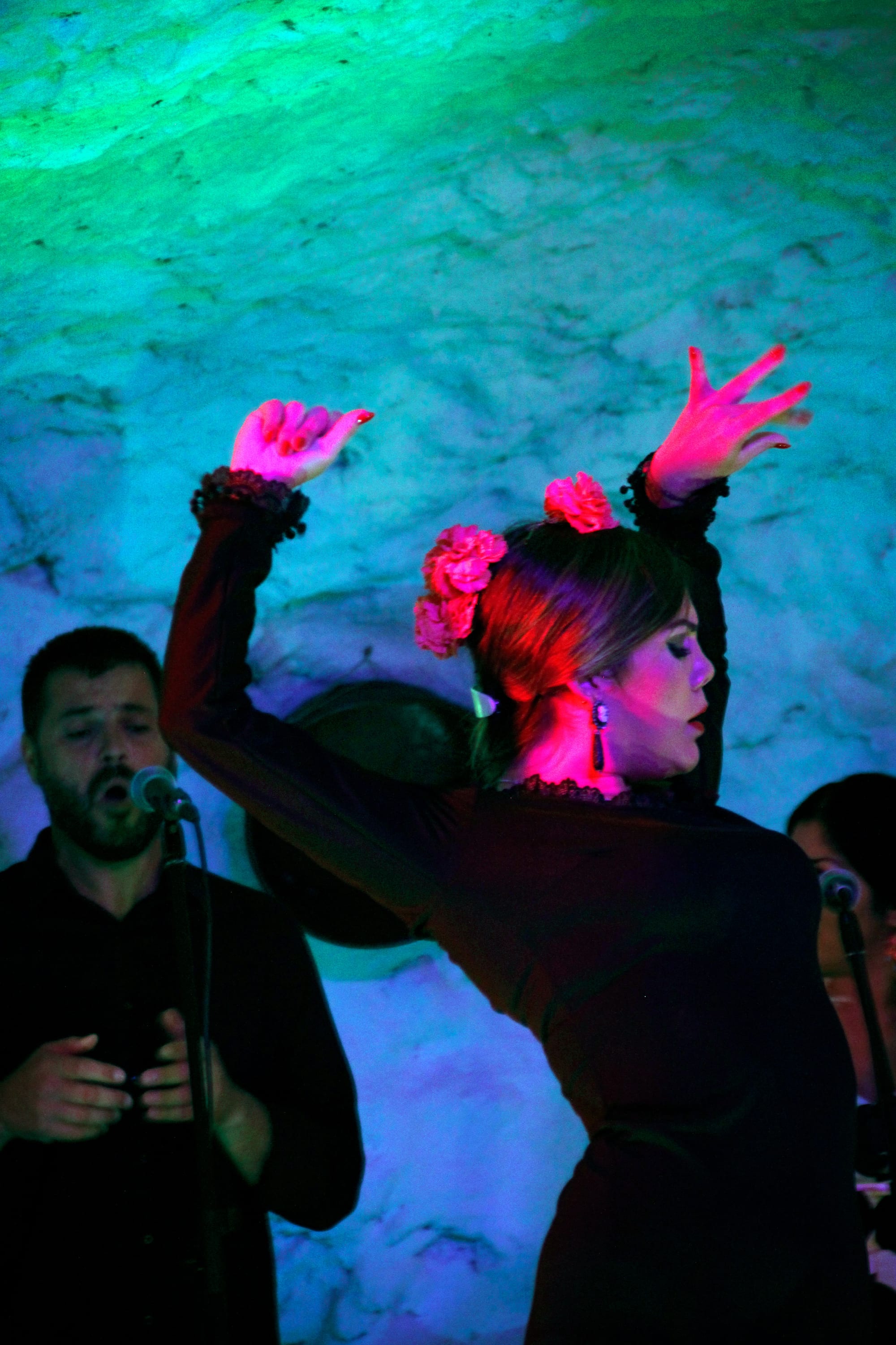 A flamenco dancer in a dark dress with pink flowers in her hair raises both arms above her head, lit in pink and green, while a male singer in black sings into a microphone beside her