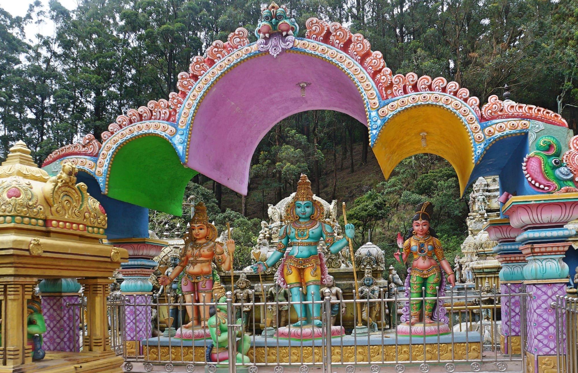 Brightly painted statues of Hindu deities in blue, gold and green stand beneath a large arch coloured pink, green, yellow and blue, with forested hillside behind