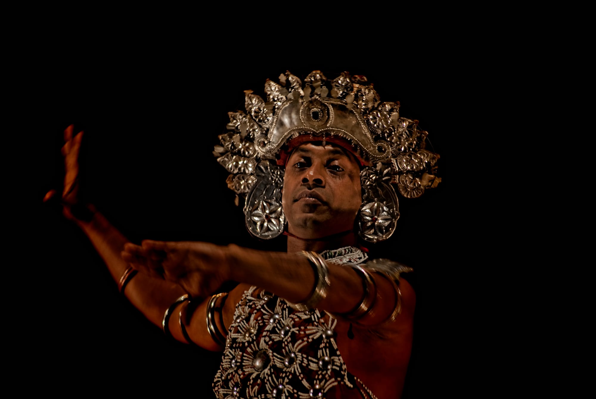 A male dancer wearing a tall ornate silver headdress, a beaded breastplate and silver bangles stretches both arms out to one side against a black background