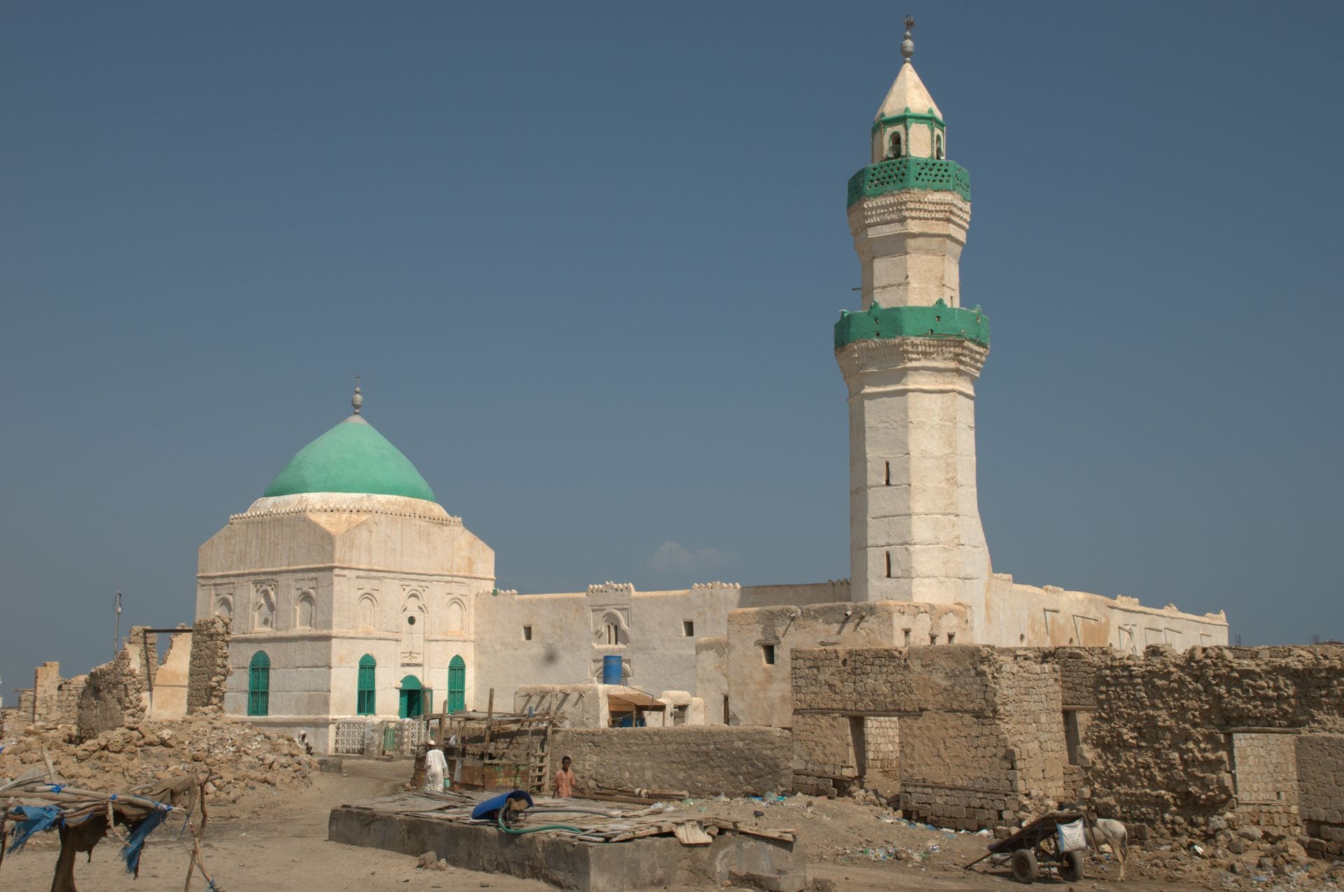A whitewashed coral-stone mosque with a green dome and minaret above a ruined old town