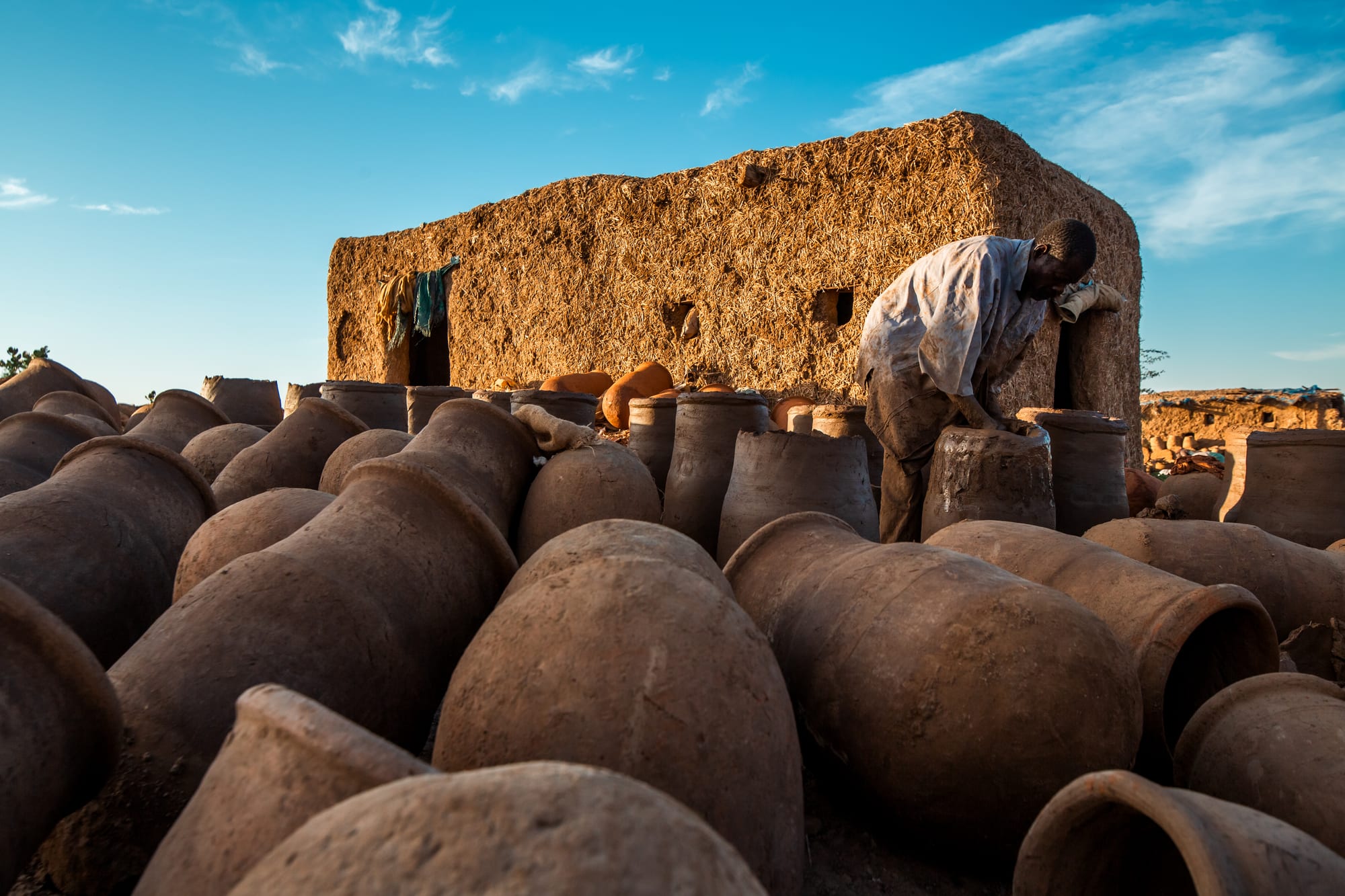 Dozens of large unglazed clay jars lie and stand in rows in the foreground while a man bends over one of them beside a mud-brick building under a blue sky