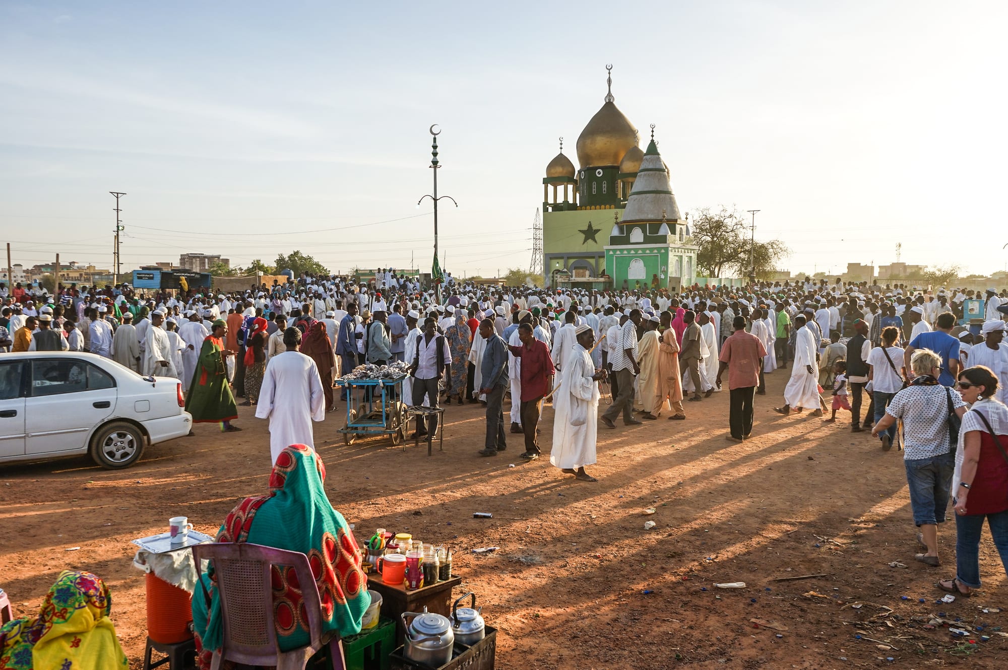 A large crowd of men, most in white robes and caps, gathers on open dusty ground in low evening sun before a green building topped with golden domes, with a woman in a bright headscarf at a tea stall in the foreground
