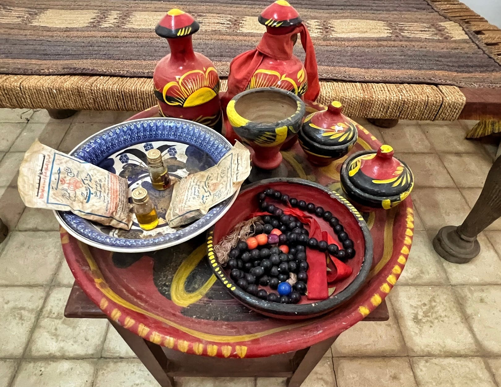 A large round red painted tray holding lidded red and yellow pots, a blue and white dish with small perfume bottles and paper packets, and a bowl of dark prayer beads tied with red ribbon