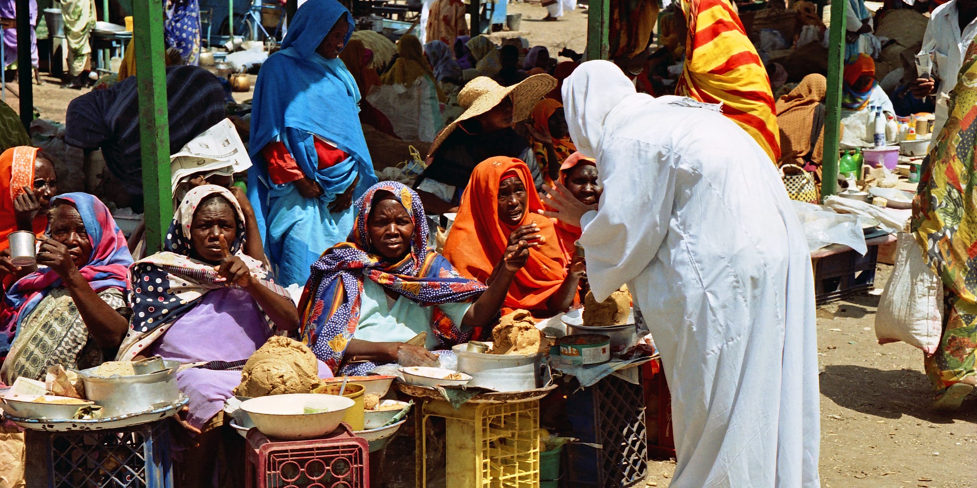 Women in colourful headscarves seated at a busy outdoor food market in Khartoum, with bowls and containers of food on low tables