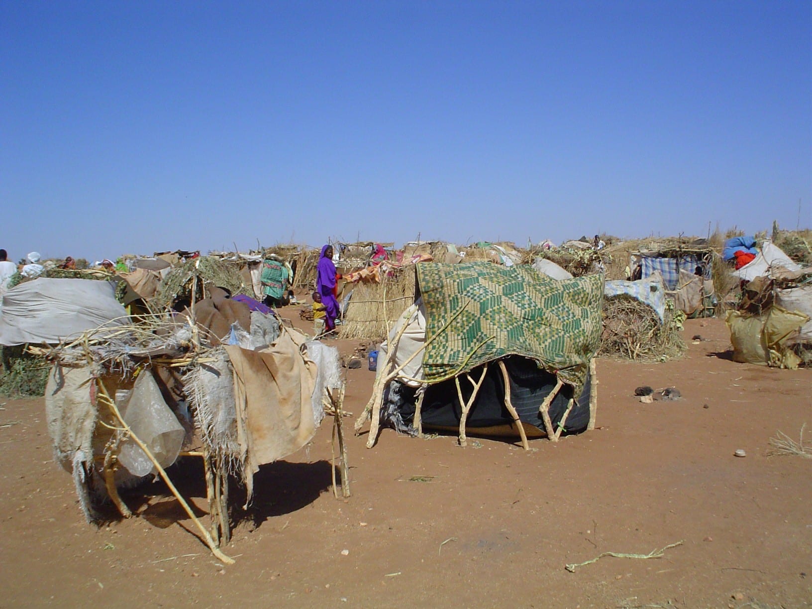 Makeshift shelters built from sticks, matting and scraps of cloth spread across a dry, open plain under a clear sky