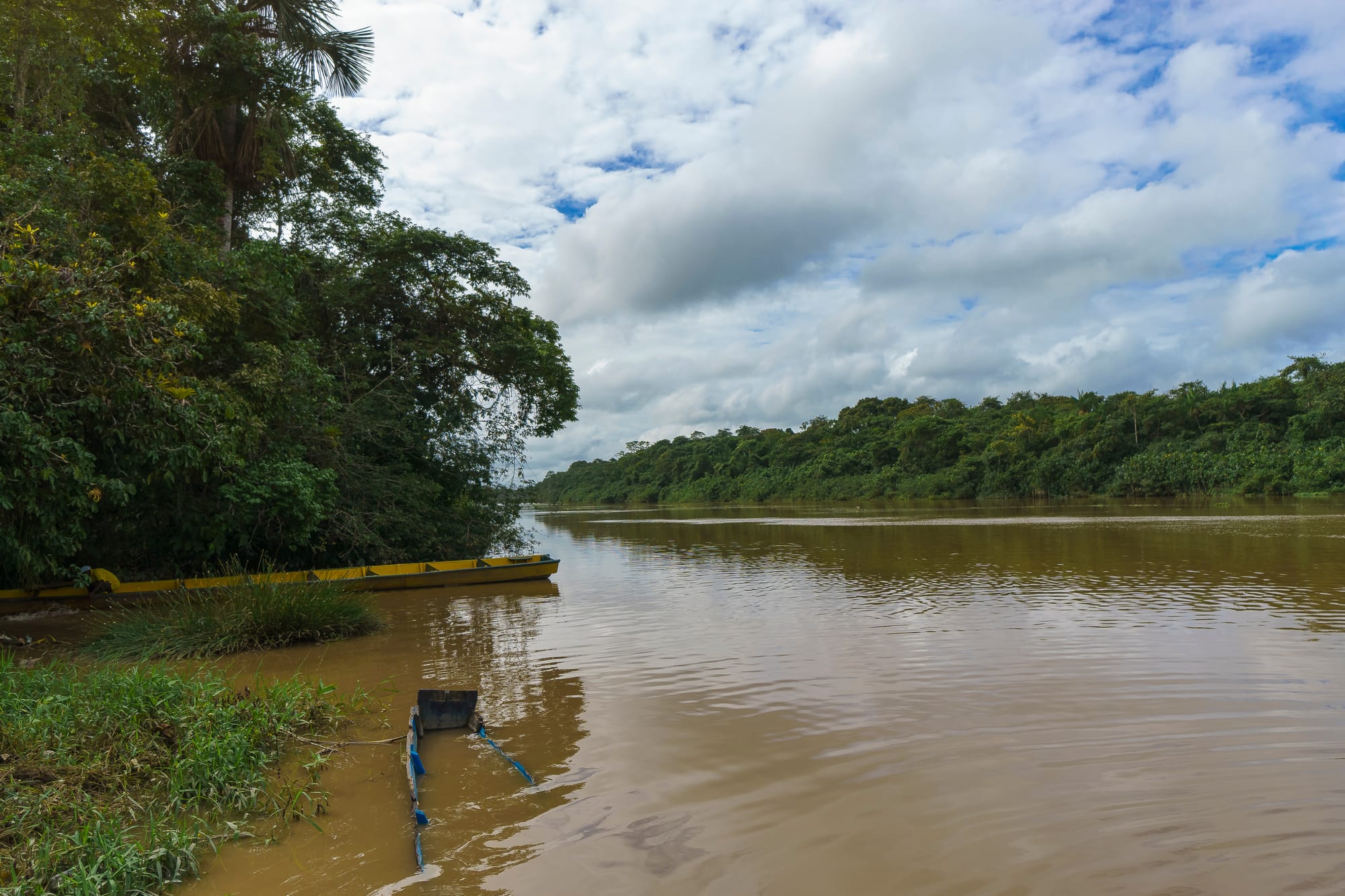 Dugout canoes moored along a wide jungle river under a partly cloudy sky