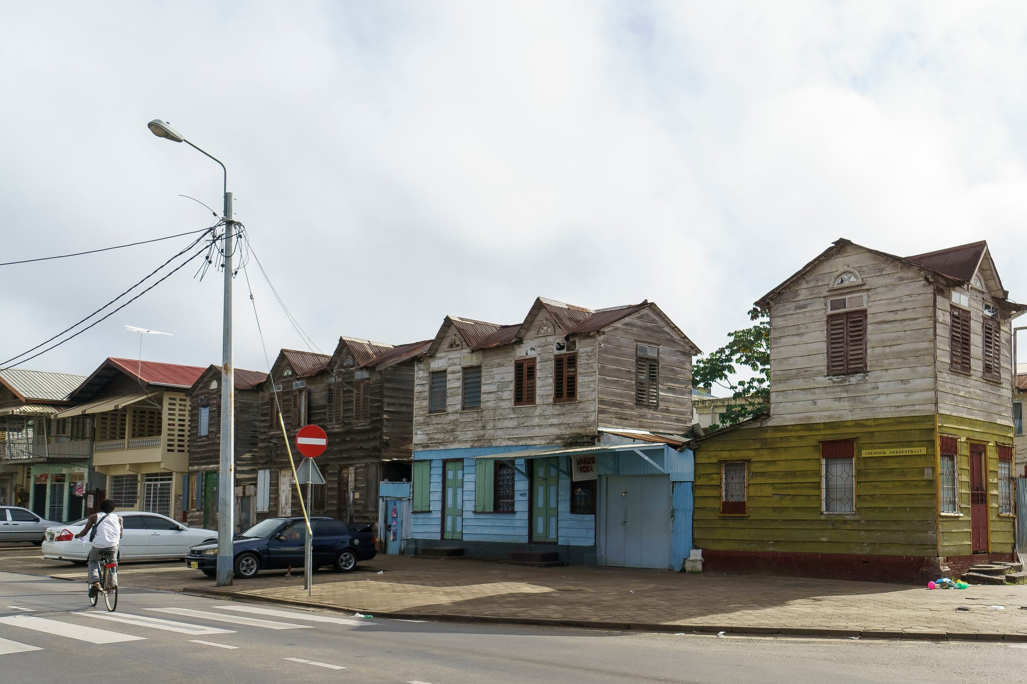 A street of weathered wooden houses in Paramaribo, with cars, a cyclist and a no-entry sign
