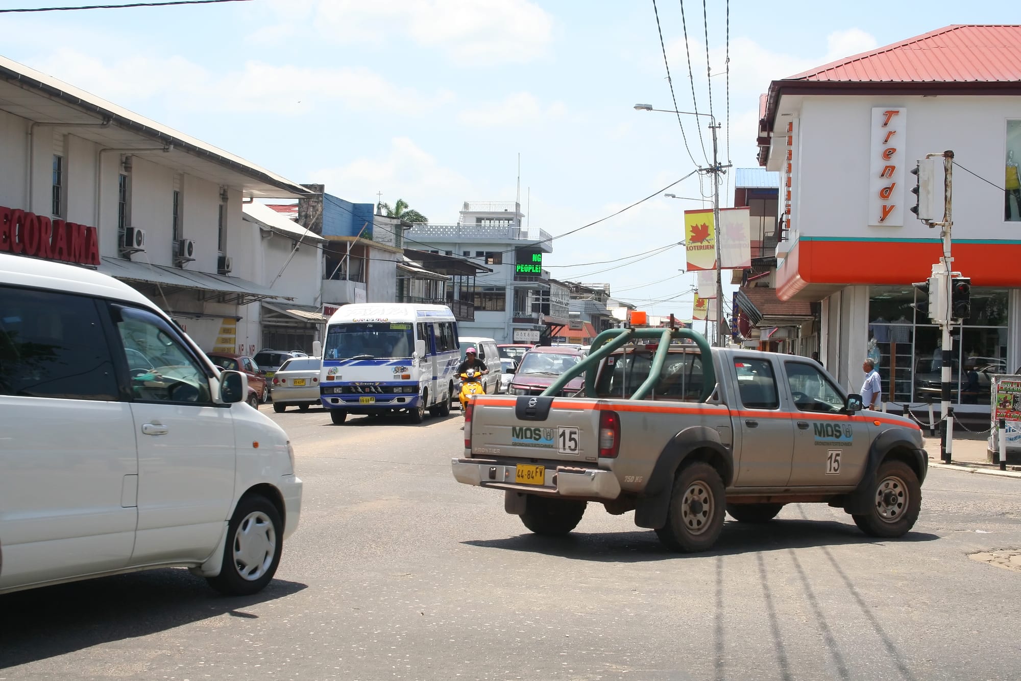 A busy Paramaribo street junction with a minibus, cars, a pickup truck and shopfronts