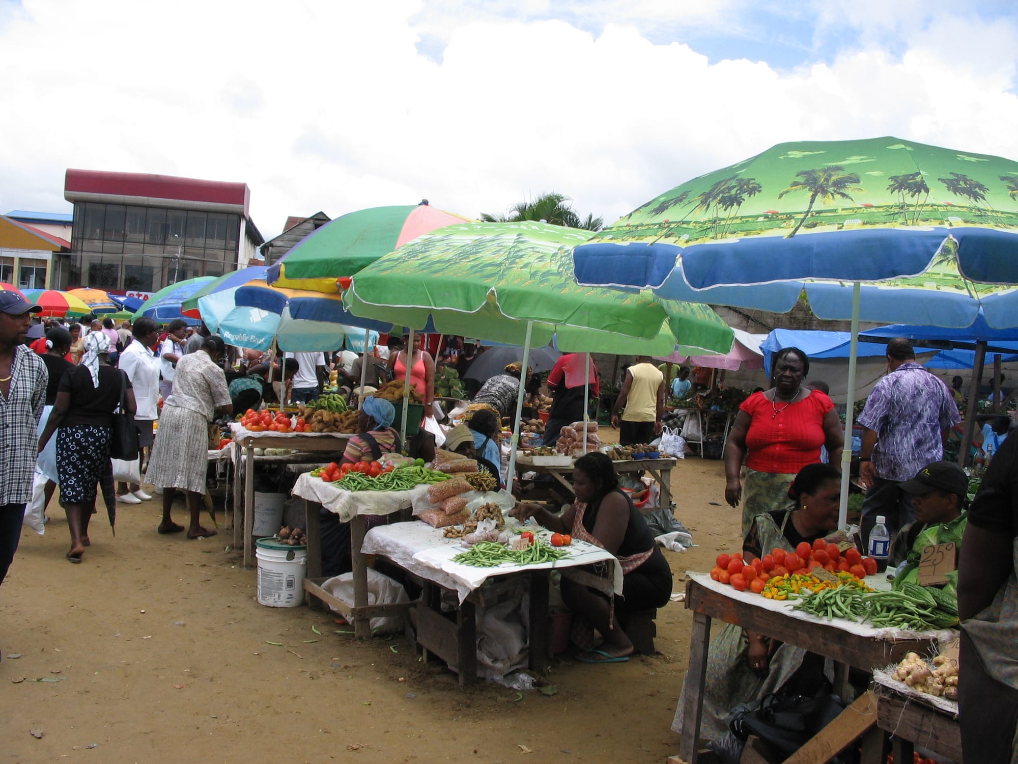 A crowded outdoor market in Paramaribo with colourful umbrellas shading stalls of tomatoes, beans and vegetables