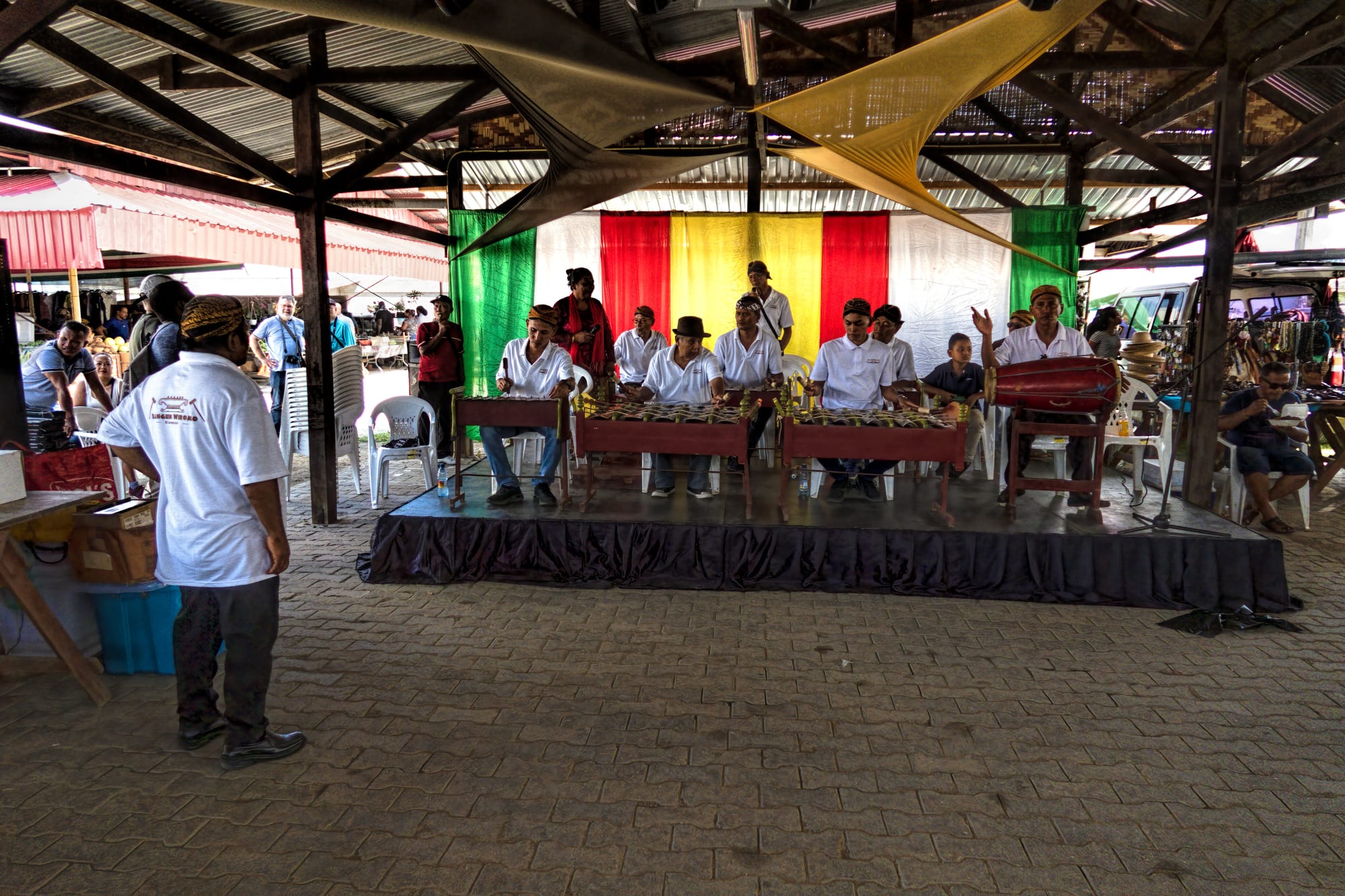 A gamelan ensemble of men in white shirts and dark caps plays metallophones and a drum on a low stage under a market roof, with a red, yellow and green backdrop