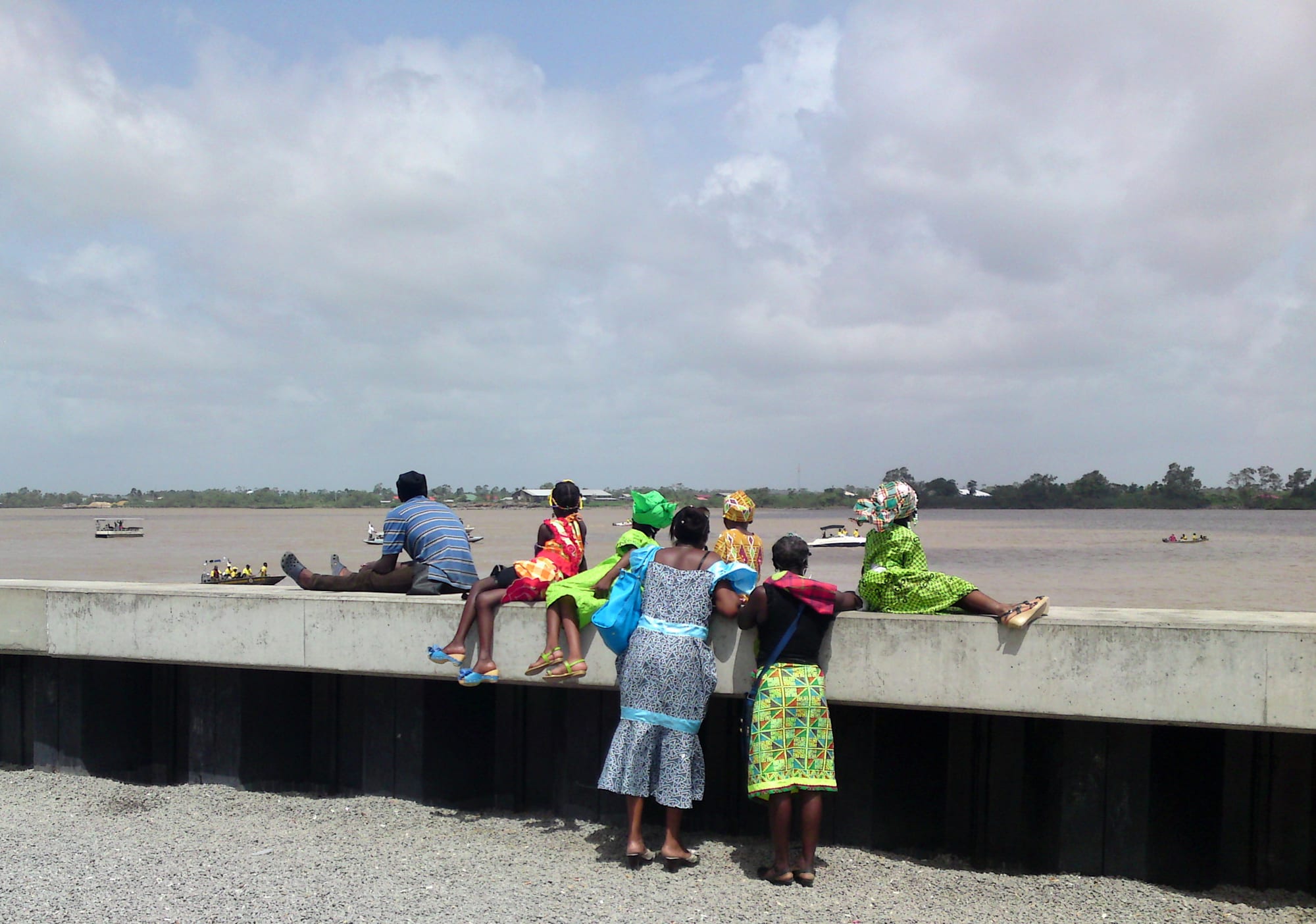 Women in brightly patterned dresses and folded headscarves stand and sit along a concrete river wall, seen from behind, looking out over a wide brown river with small boats