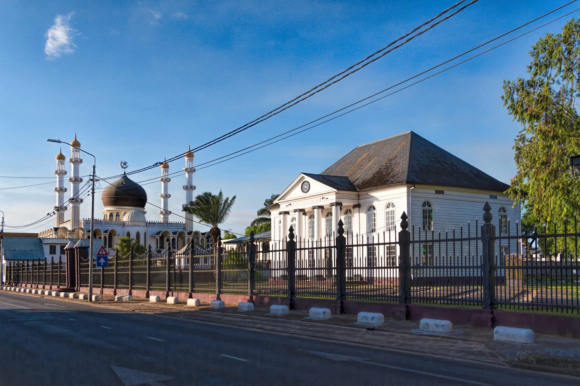 A white wooden synagogue with a columned portico stands behind iron railings, with a large mosque with a dark dome and four slender white minarets directly beside it under a blue sky