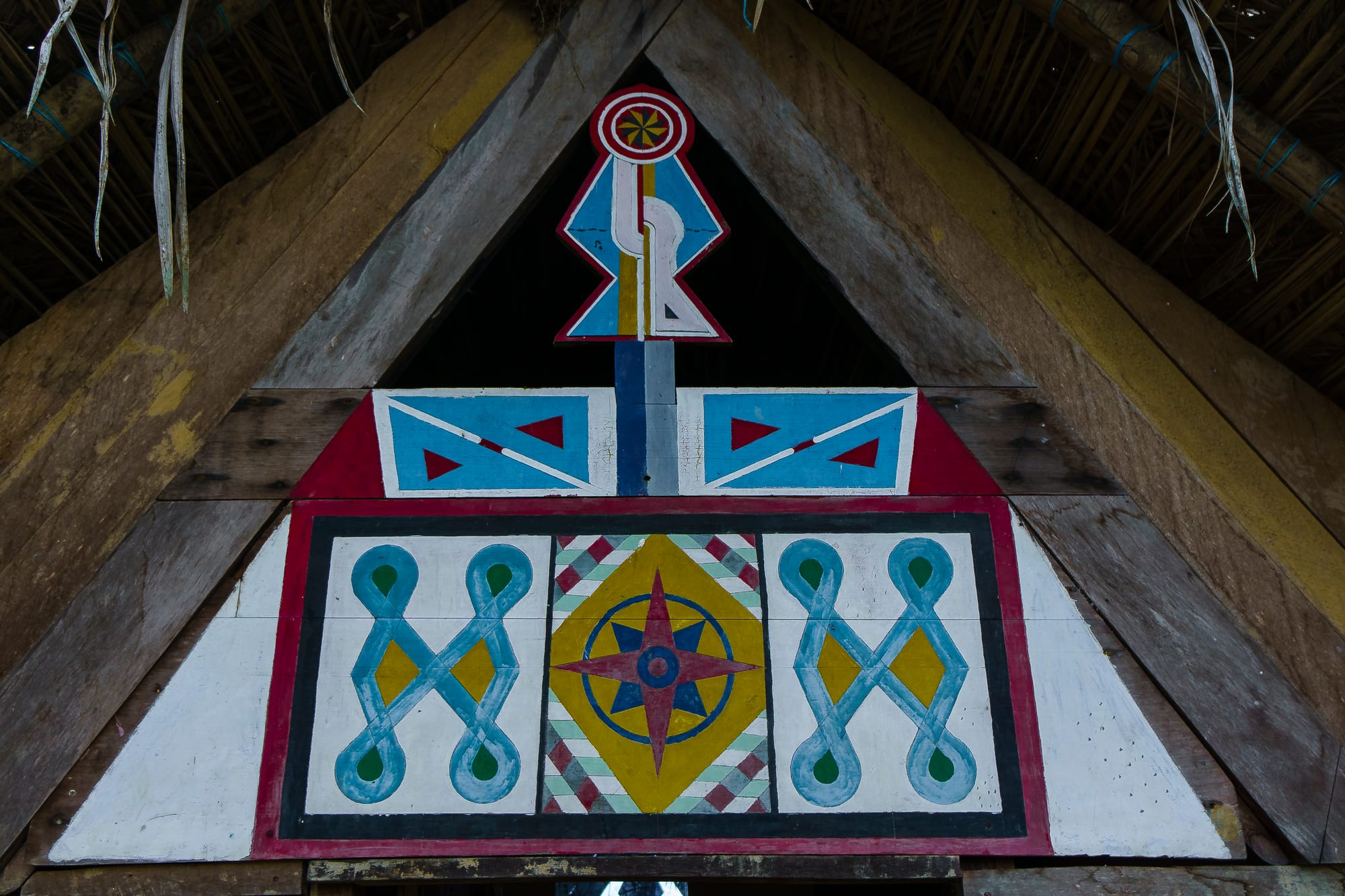 The triangular gable of a thatched wooden building painted with symmetrical geometric Maroon designs in red, blue, yellow and white, including interlaced loops and a central star