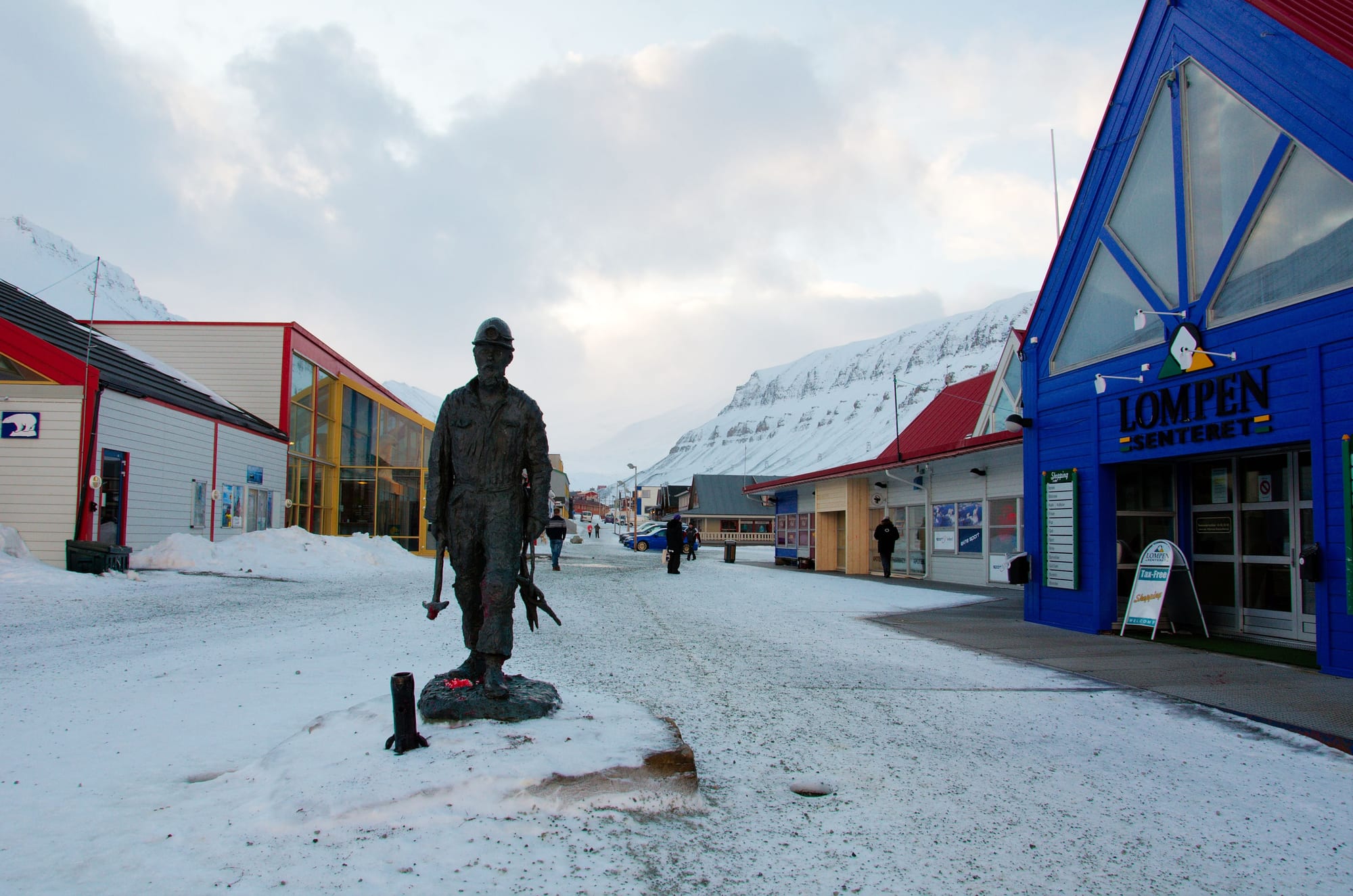 A snow-covered pedestrian street in Longyearbyen lined with colourful shopfronts and a statue of a miner, mountains behind