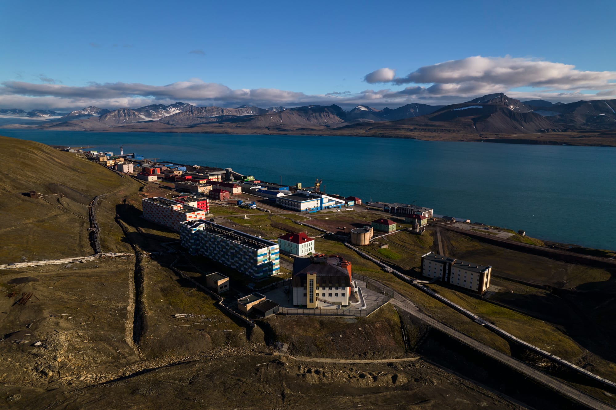 An aerial view of a small settlement of apartment blocks and industrial buildings on a treeless slope beside a wide fjord, with snow-streaked mountains beyond