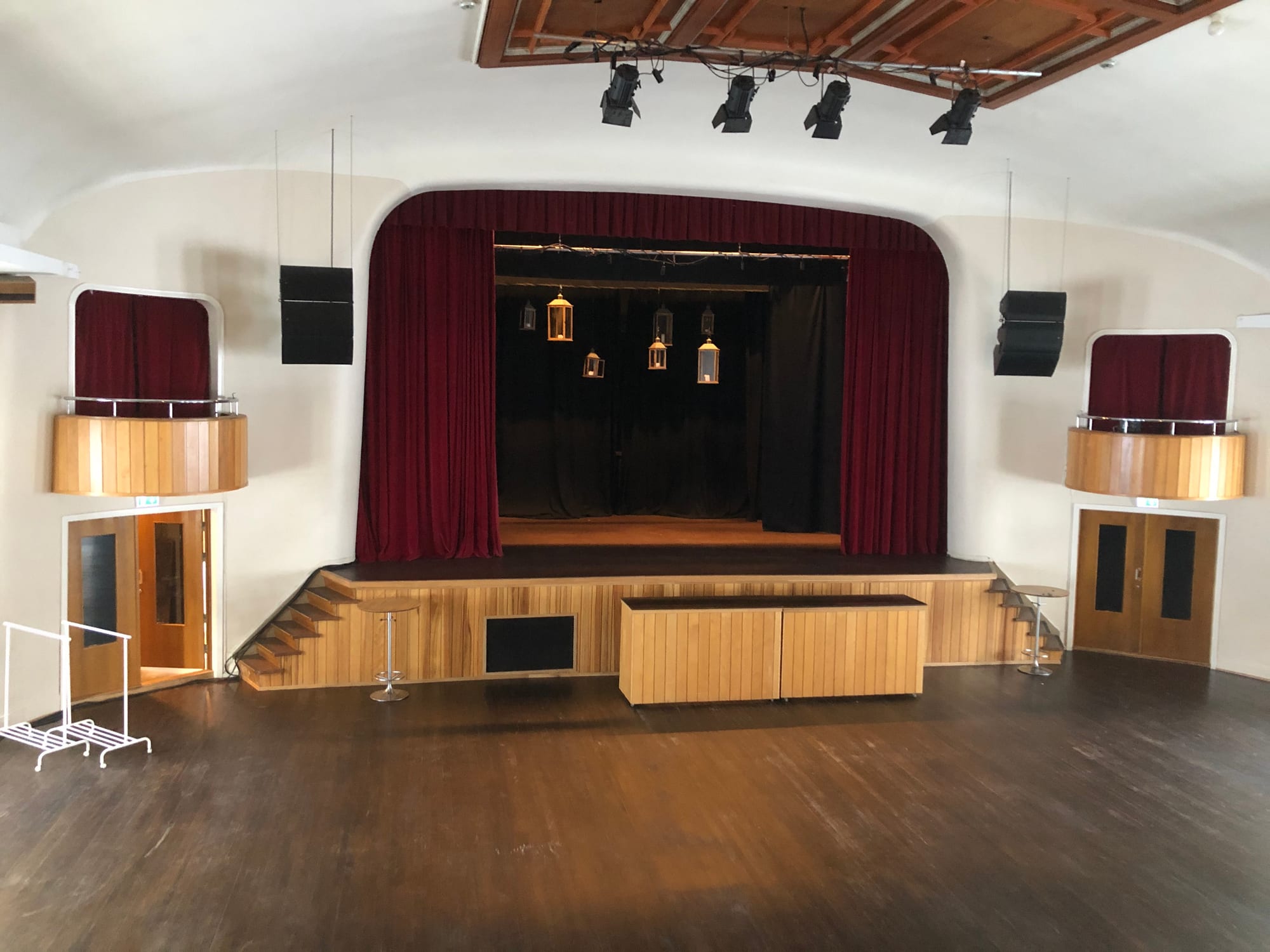 An empty hall with a dark wooden floor, a stage framed by deep red curtains with lanterns hanging inside, stage lights on the ceiling and small curtained balconies on each side