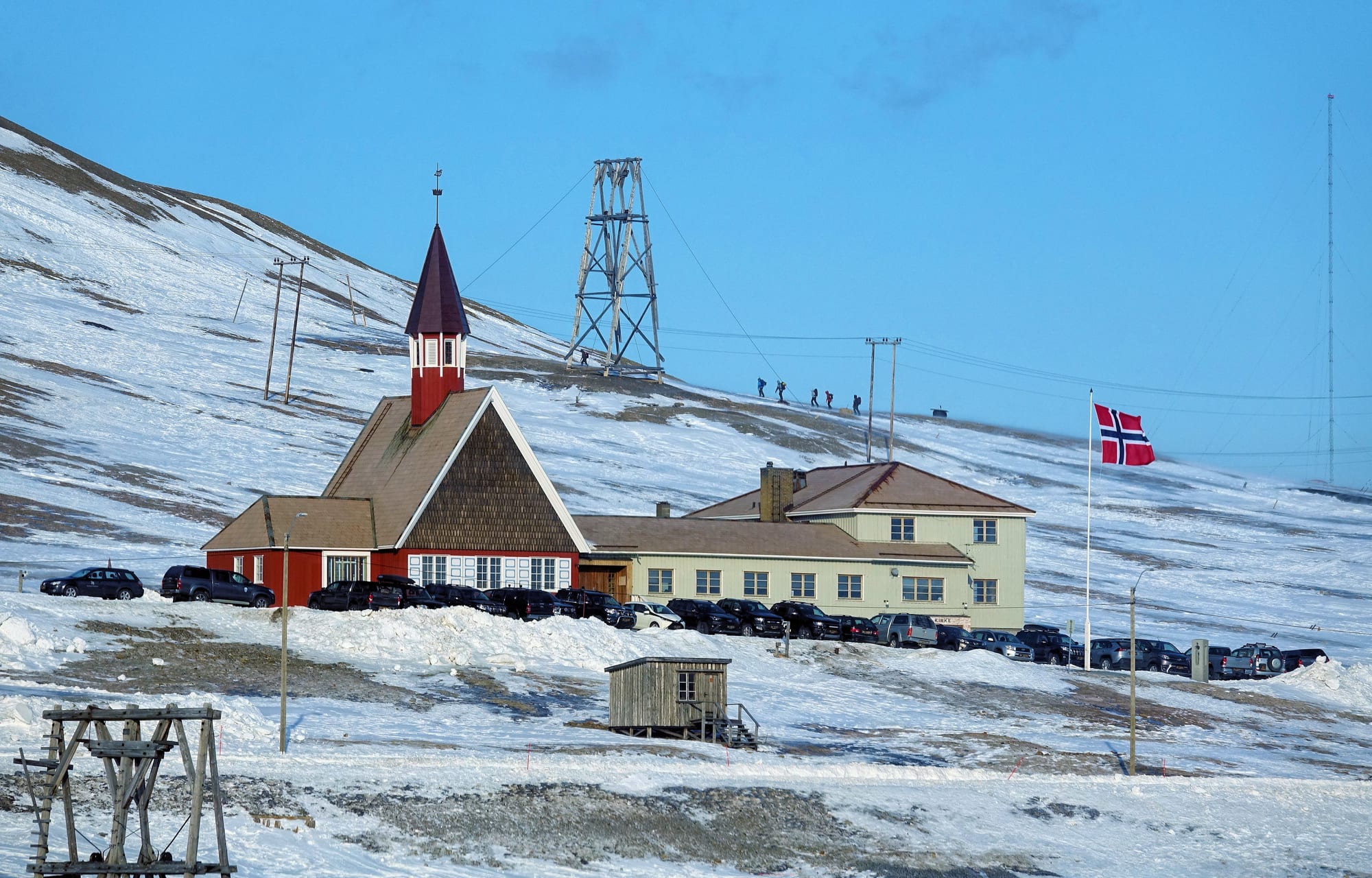 A red wooden church with a pointed spire and a pale yellow parish house stand on a snowy slope, with cars parked outside, a Norwegian flag flying and an old mining cableway tower on the hillside behind