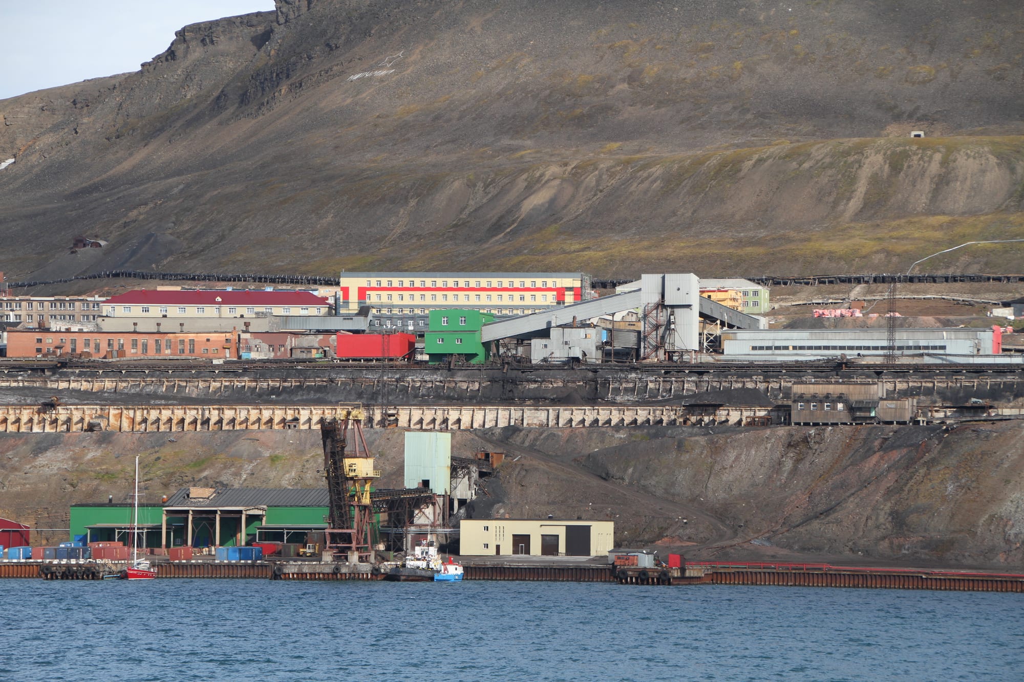 Colourful apartment blocks and coal-handling buildings rise in tiers above a quay on a fjord, below a bare brown mountainside